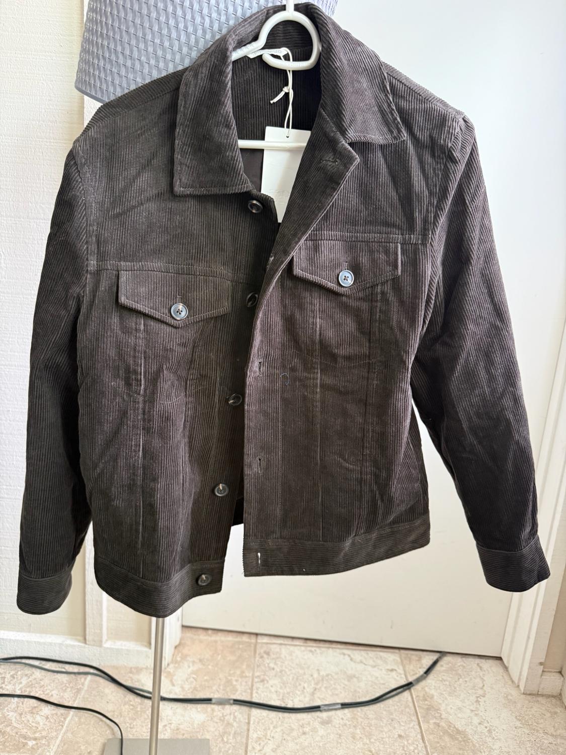 Dark brown corduroy jacket with label, buttons and front pockets, hanging on a white hanger on a metal stand.