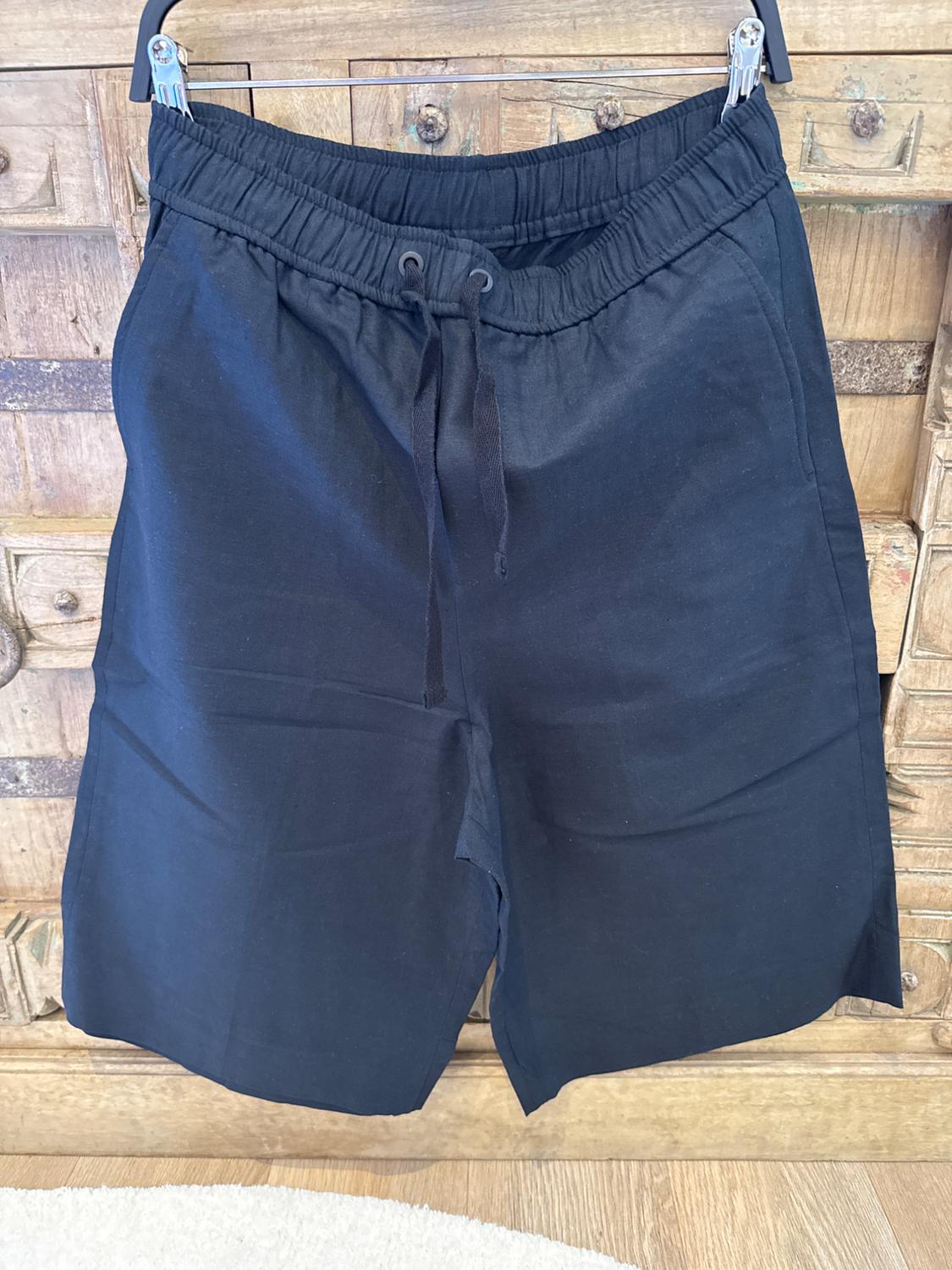 Black shorts hanging on a hanger against a wooden background. Elasticated waist with adjustable drawstring.