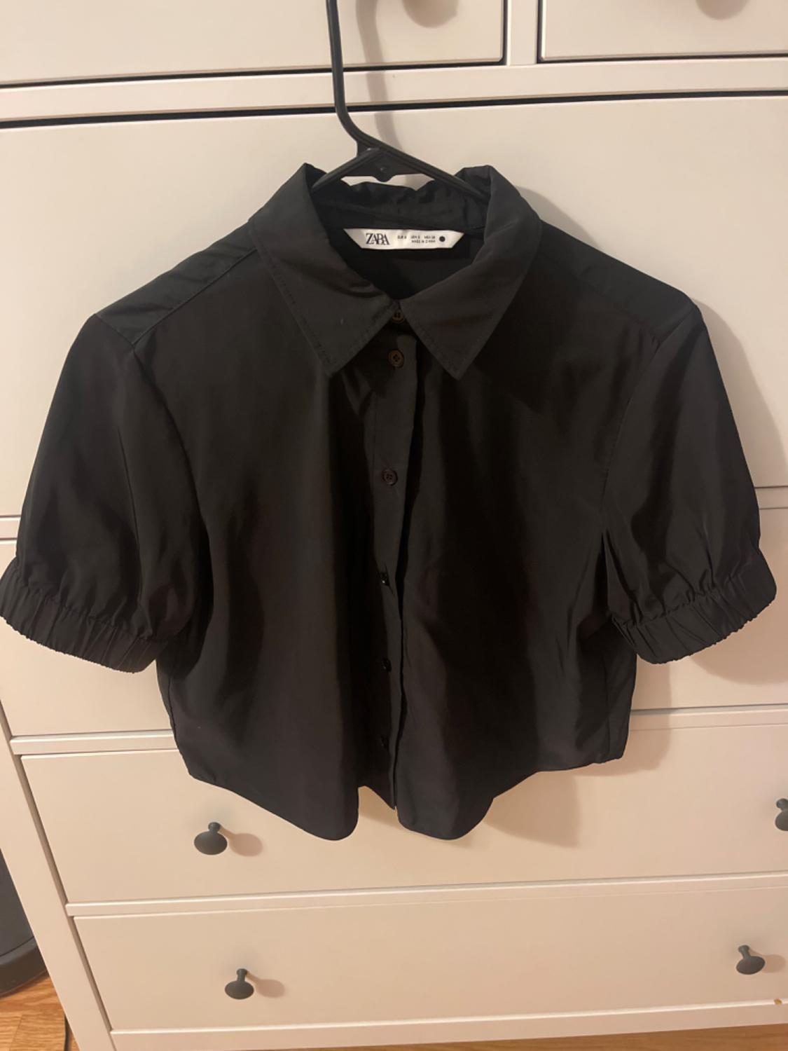 Front view of a black blouse with a classic collar and short puffed sleeves, central button fastening.