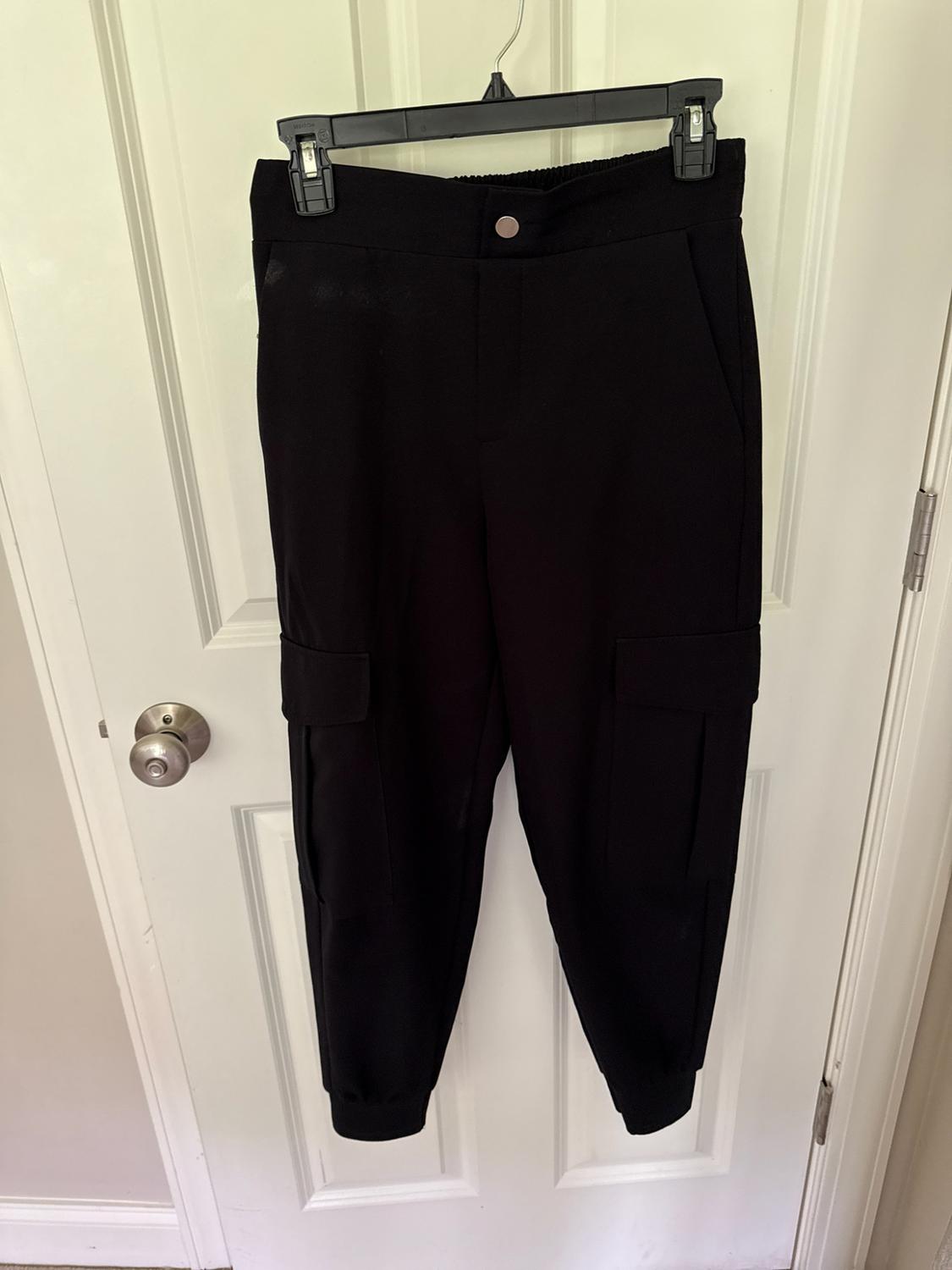 Black cargo pants hanging on a white door. Jogger design with a button closure, side flap pockets, and elastic cuffs.