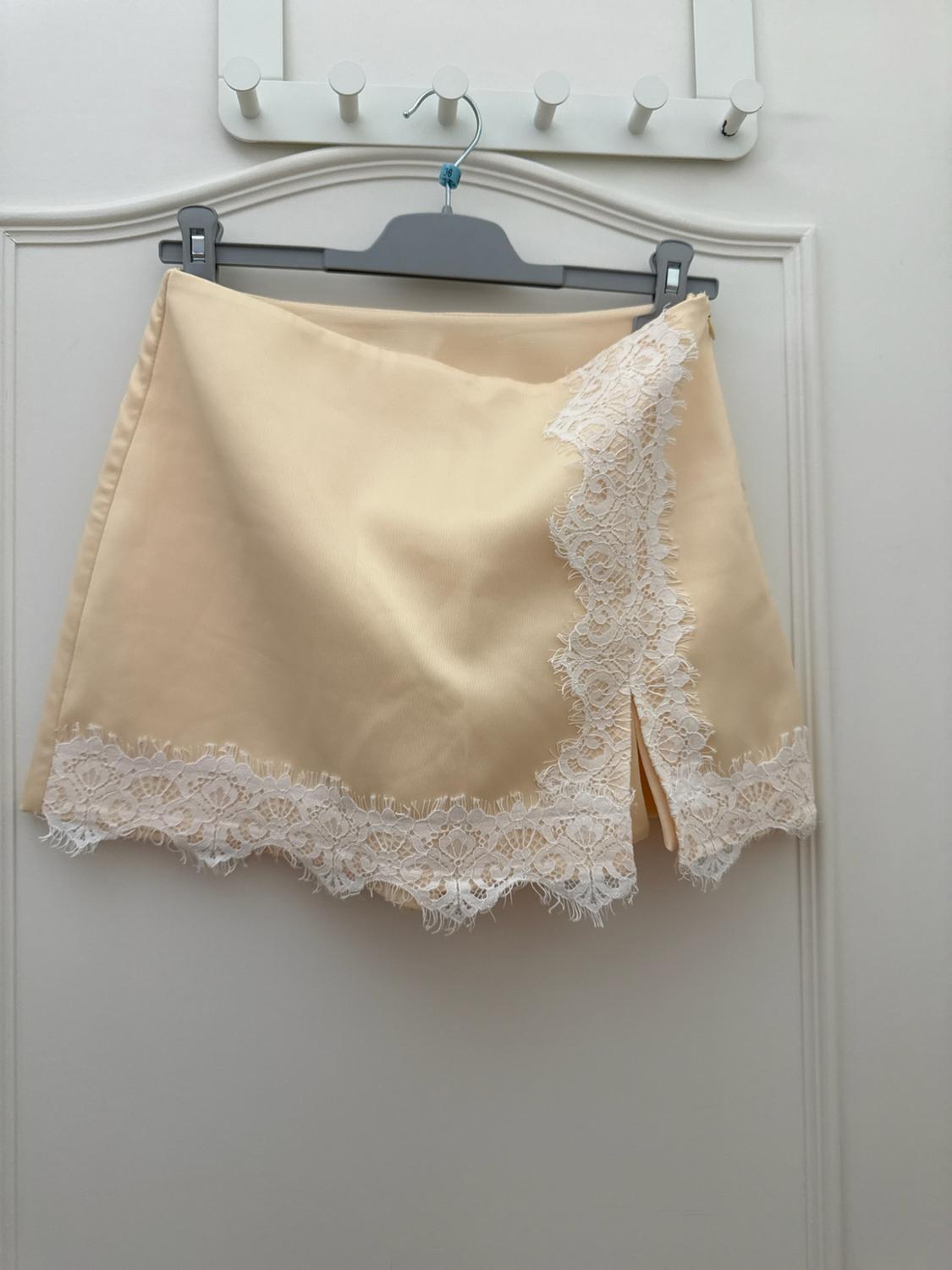 Pastel yellow mini skirt with white lace trim and side slit, hanging on a hanger over a white door.