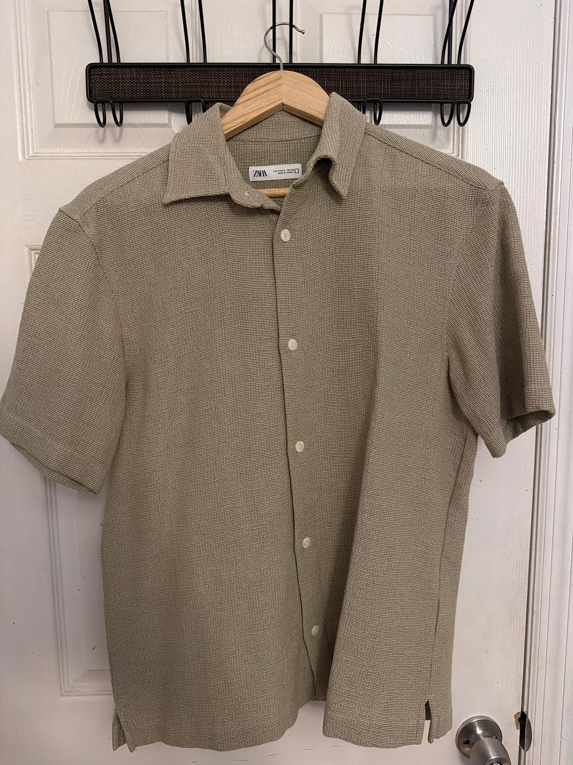 Beige short-sleeved shirt hanging on a wooden hanger, with white buttons and a light texture, over a white door.