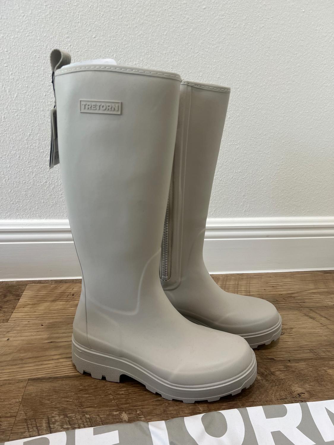 Light beige rubber tall boots on a wooden floor. Visible tag with the "TRETORN" brand. Part of a box in the foreground.
