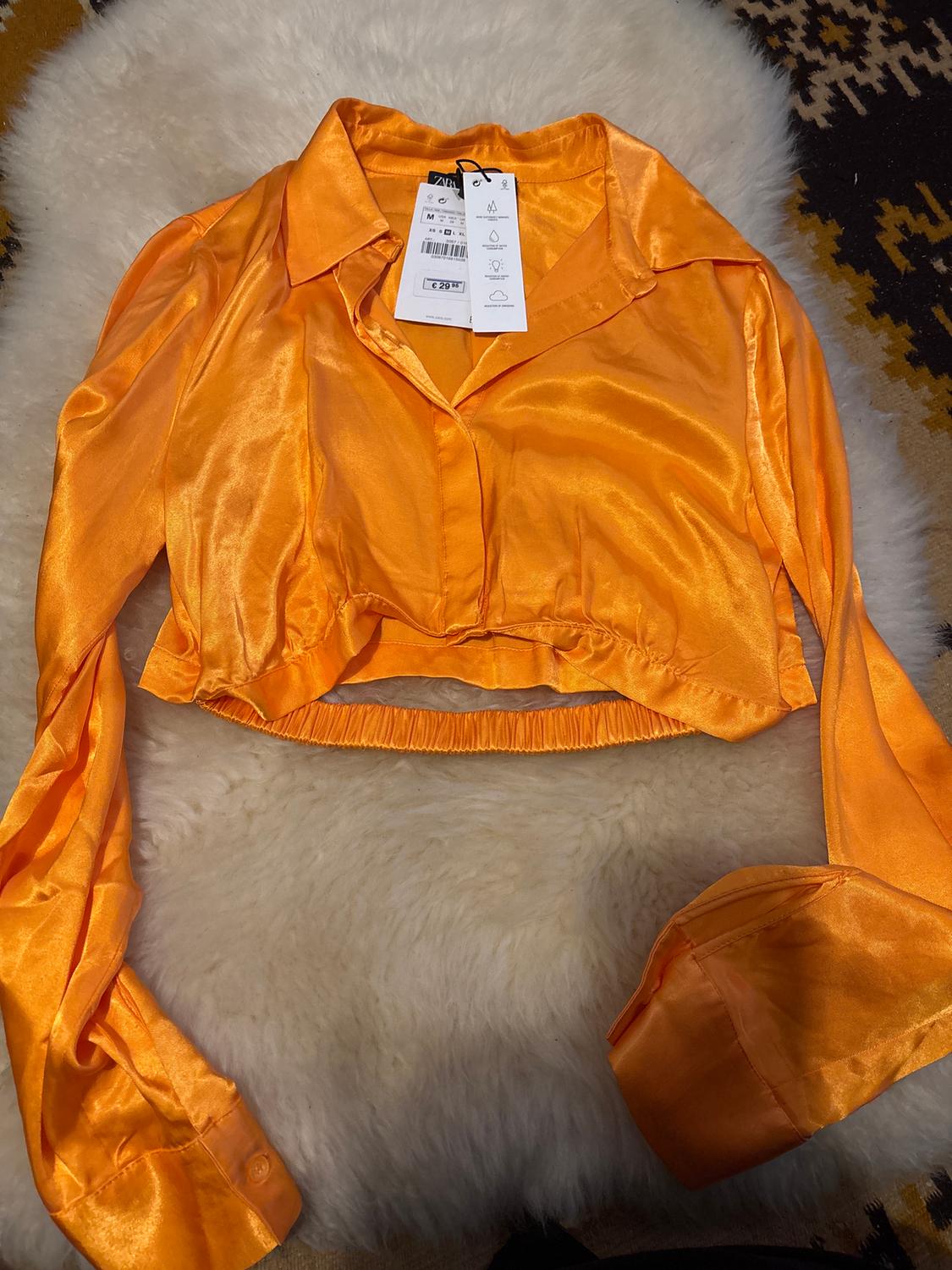 Orange satin cropped shirt with long sleeves and a classic collar, on a white rug. Visible label with care details.