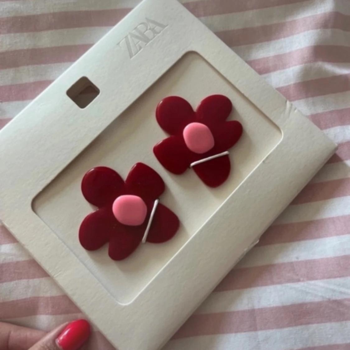 Red flower-design earrings with a pink centre, presented on card featuring the text ZARA. Someone with red nails is holding them against a pink and white striped background.