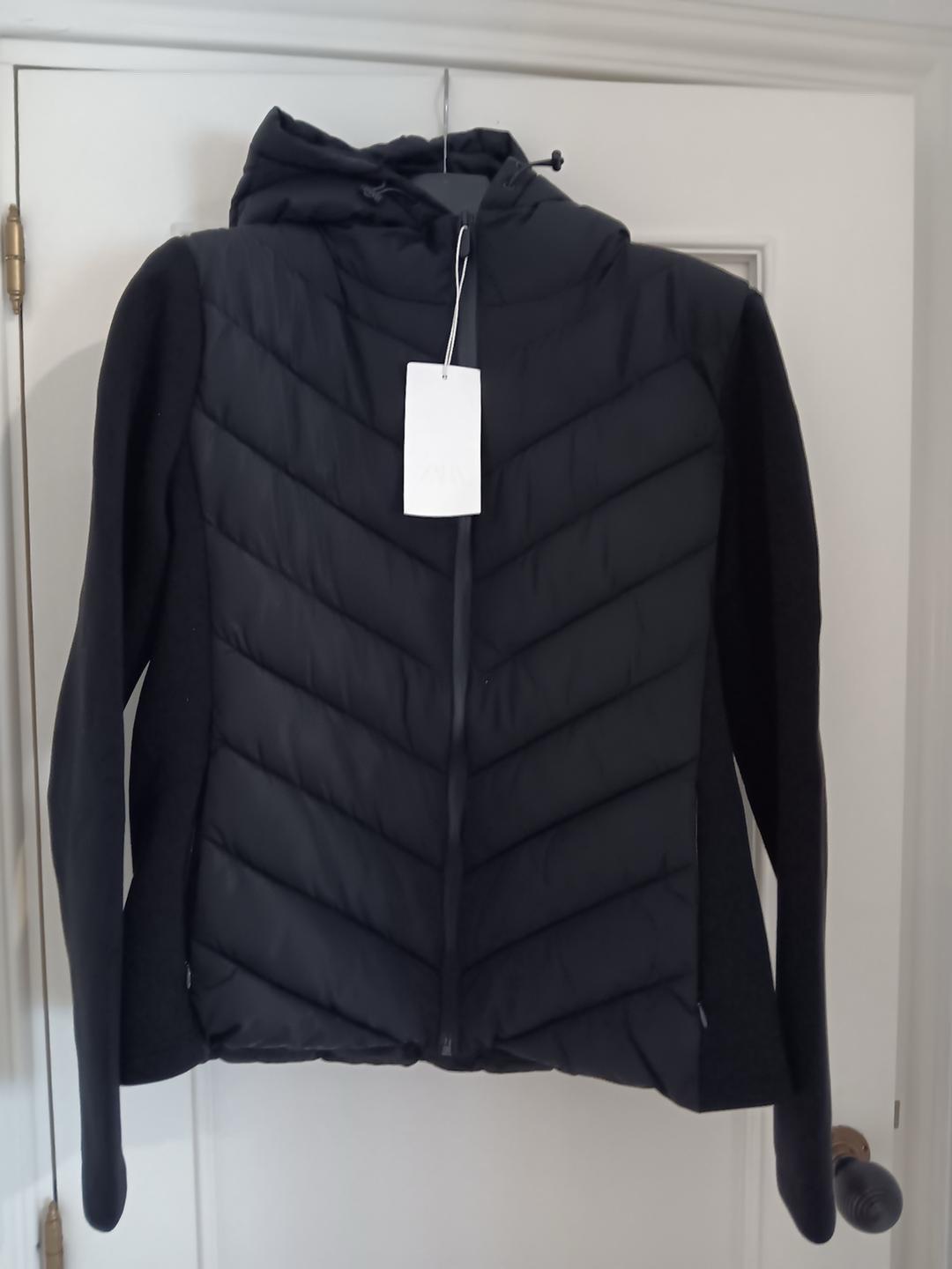 Black hooded jacket hanging on a white door. V-shaped padded front, smooth sleeves and zip. White label visible.