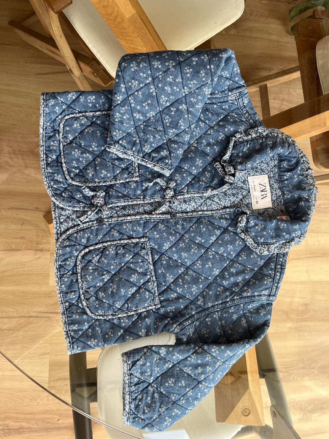 Blue padded jacket with white floral print on a wooden table. Visible label: "ZARA".