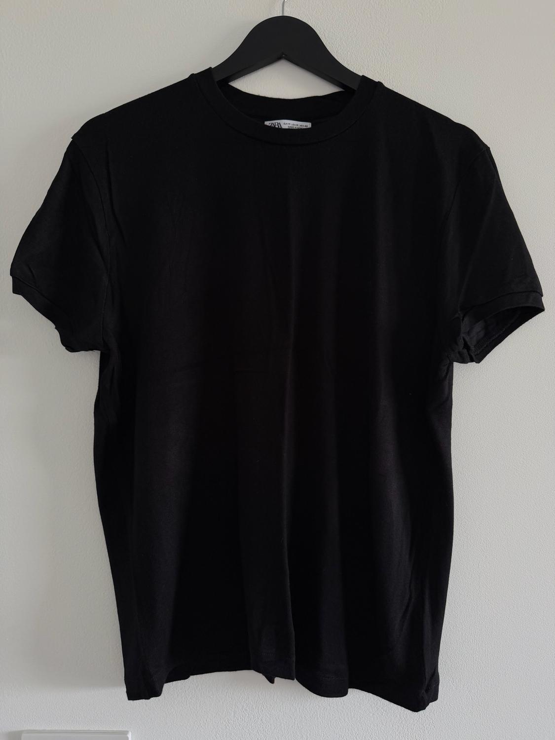 Black short-sleeved T-shirt hanging on a black hanger against a white wall.