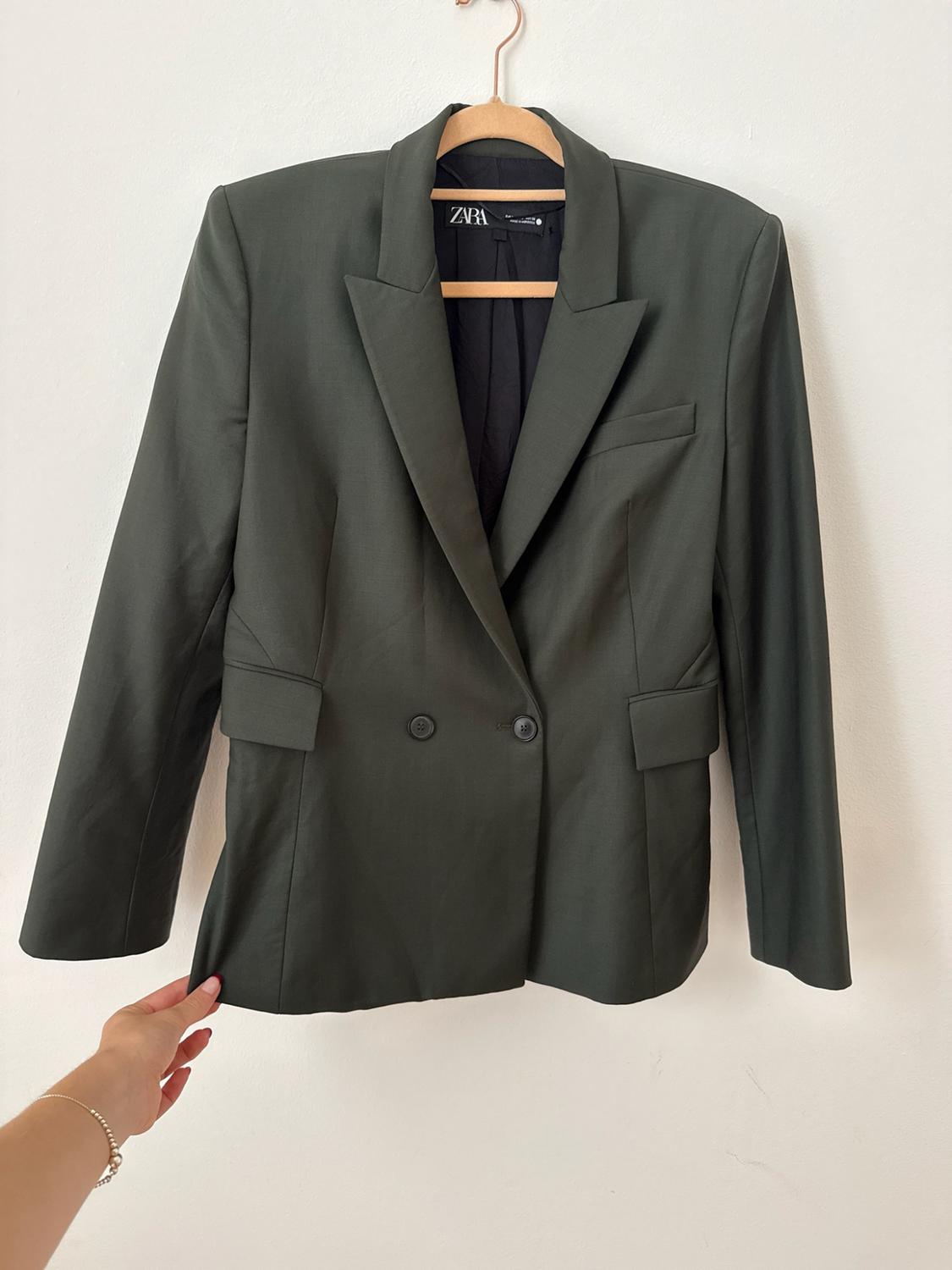 A dark green blazer is shown hanging on a wooden hanger against a white background. A hand can be seen holding the bottom edge of the garment, which includes a label with the word ZARA.