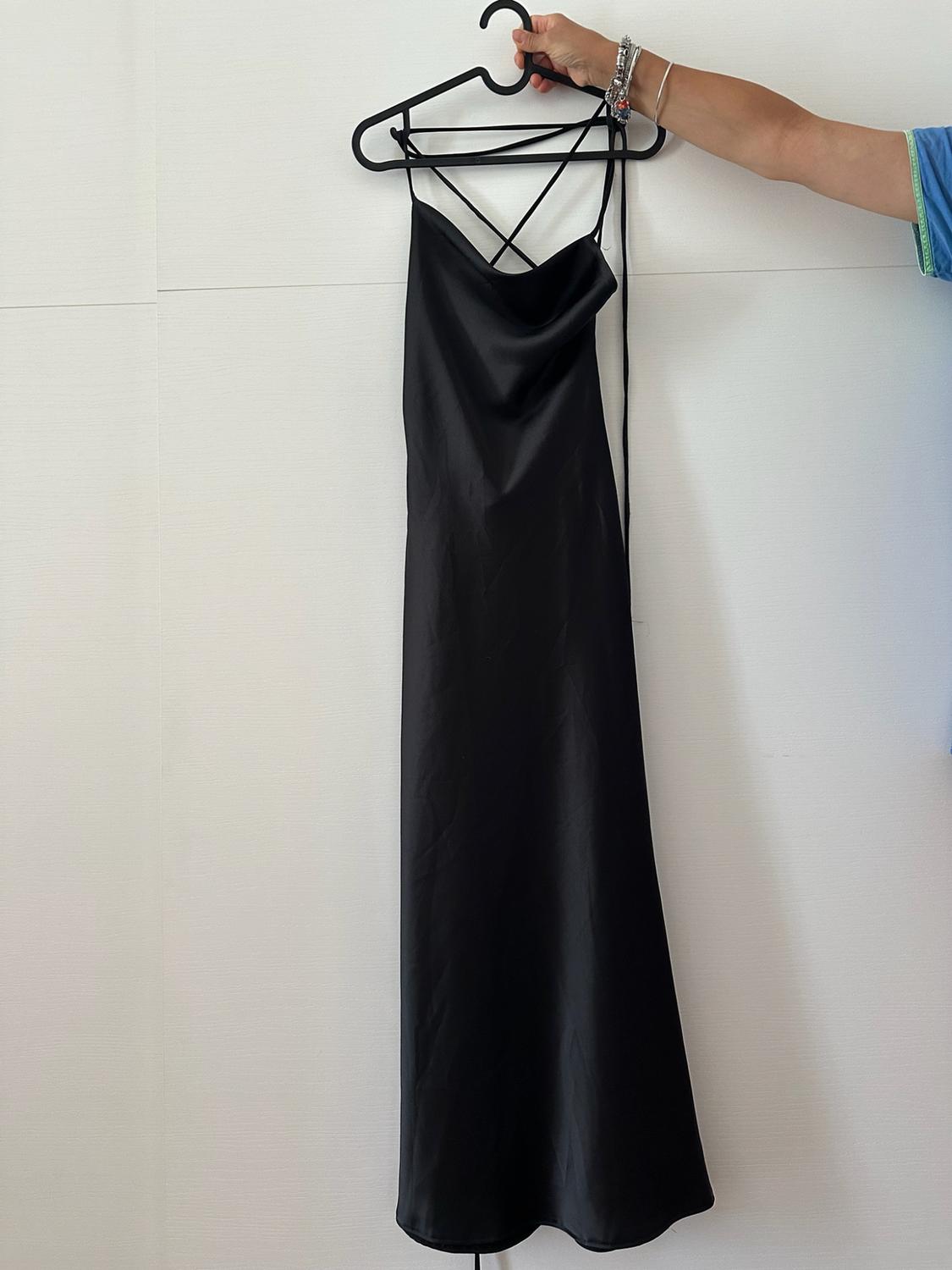 An arm with bracelets holds a hanger with a long black dress with thin crossed straps in front of a white wall.
