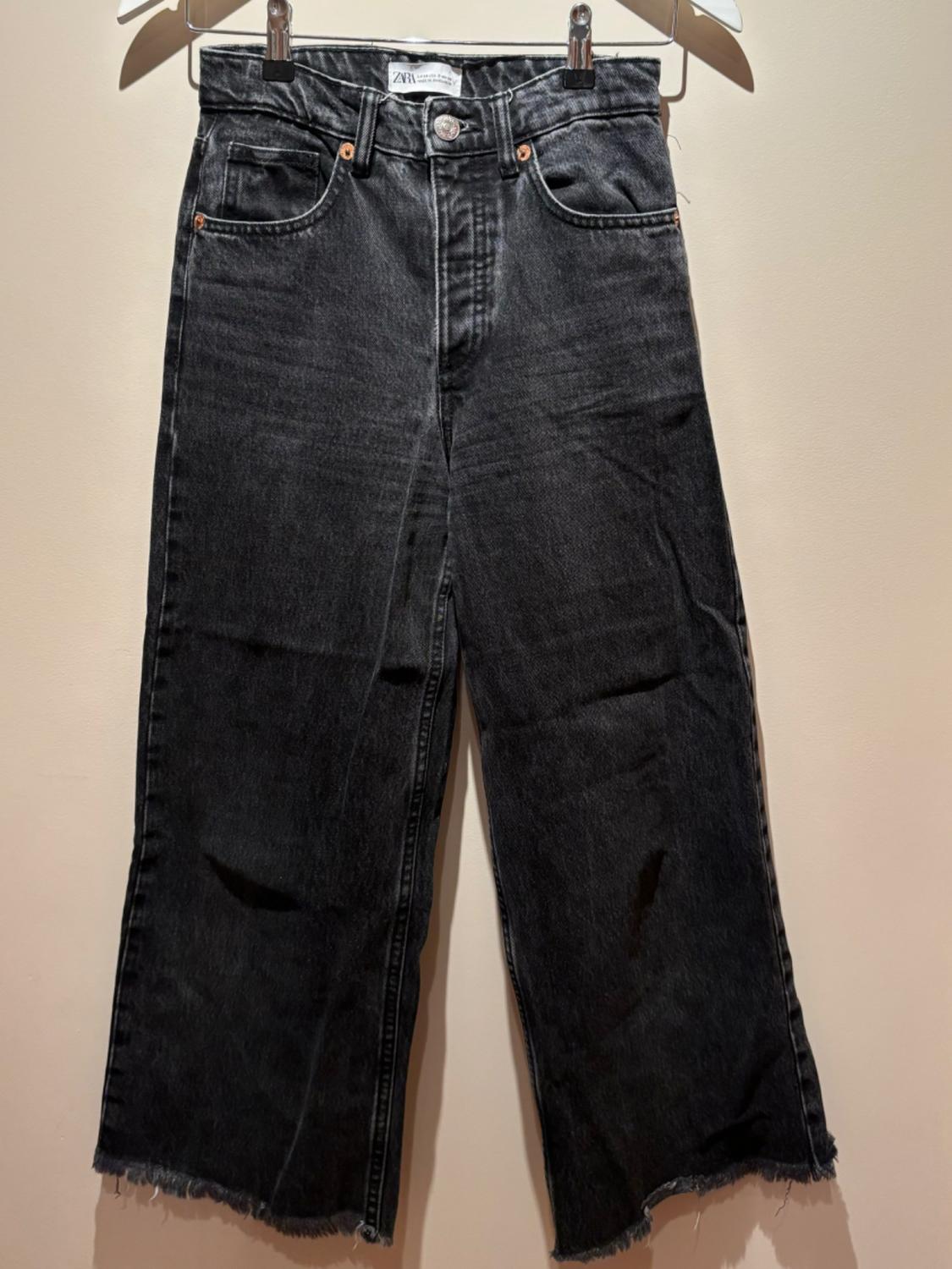 Black denim jeans with a straight leg, frayed hem detailing, hanging on a hanger against a beige background.