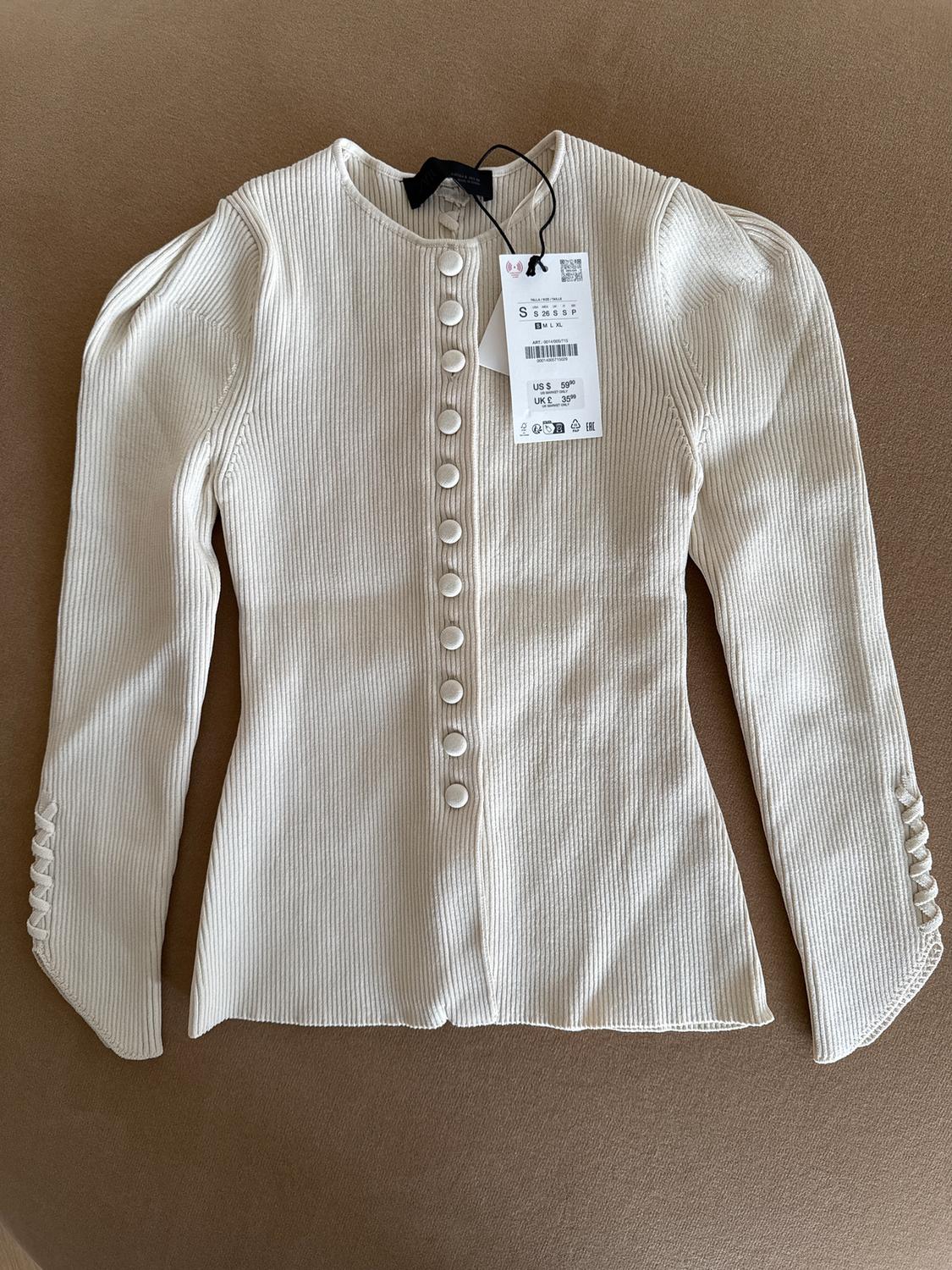 White ribbed knit jacket with front buttons and visible price tag on a brown background.