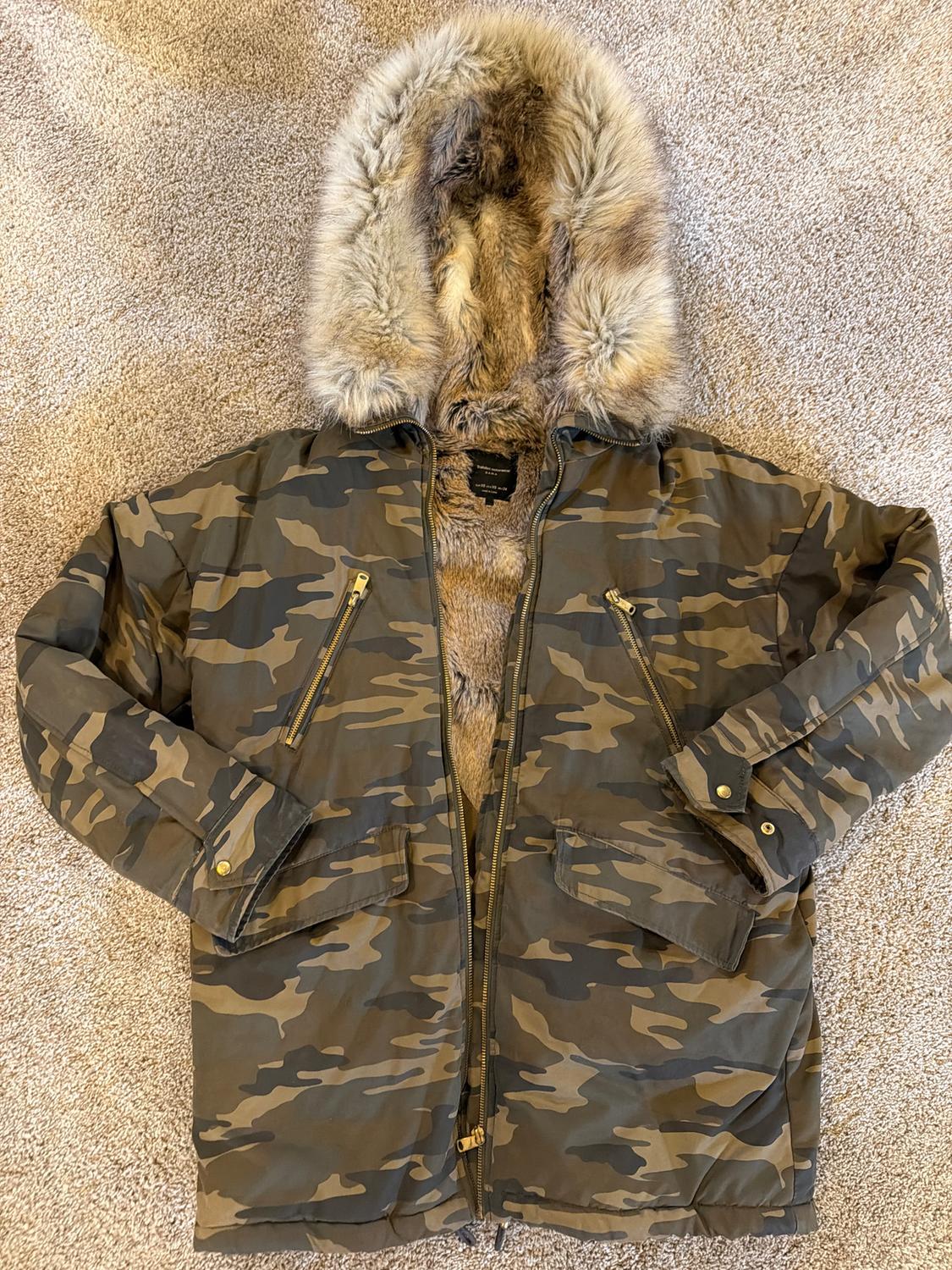Camouflage jacket with faux fur hood on a beige rug. Pockets with gold zips and furry inner lining.