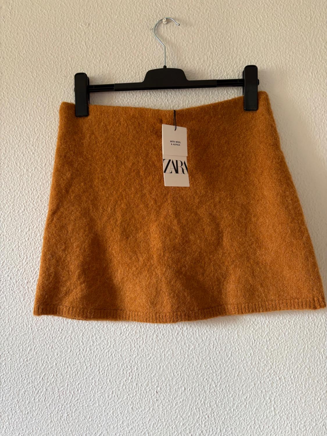 Mustard knit mini skirt on a black hanger against a white textured wall. Label: WITH WOOL & ALPACA ZARA.