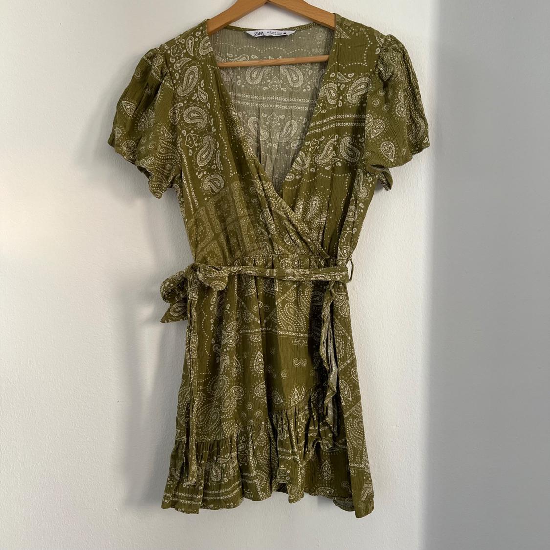 Olive green mini dress with white paisley print, short sleeves and belt, hanging on a wooden hanger against a white wall.