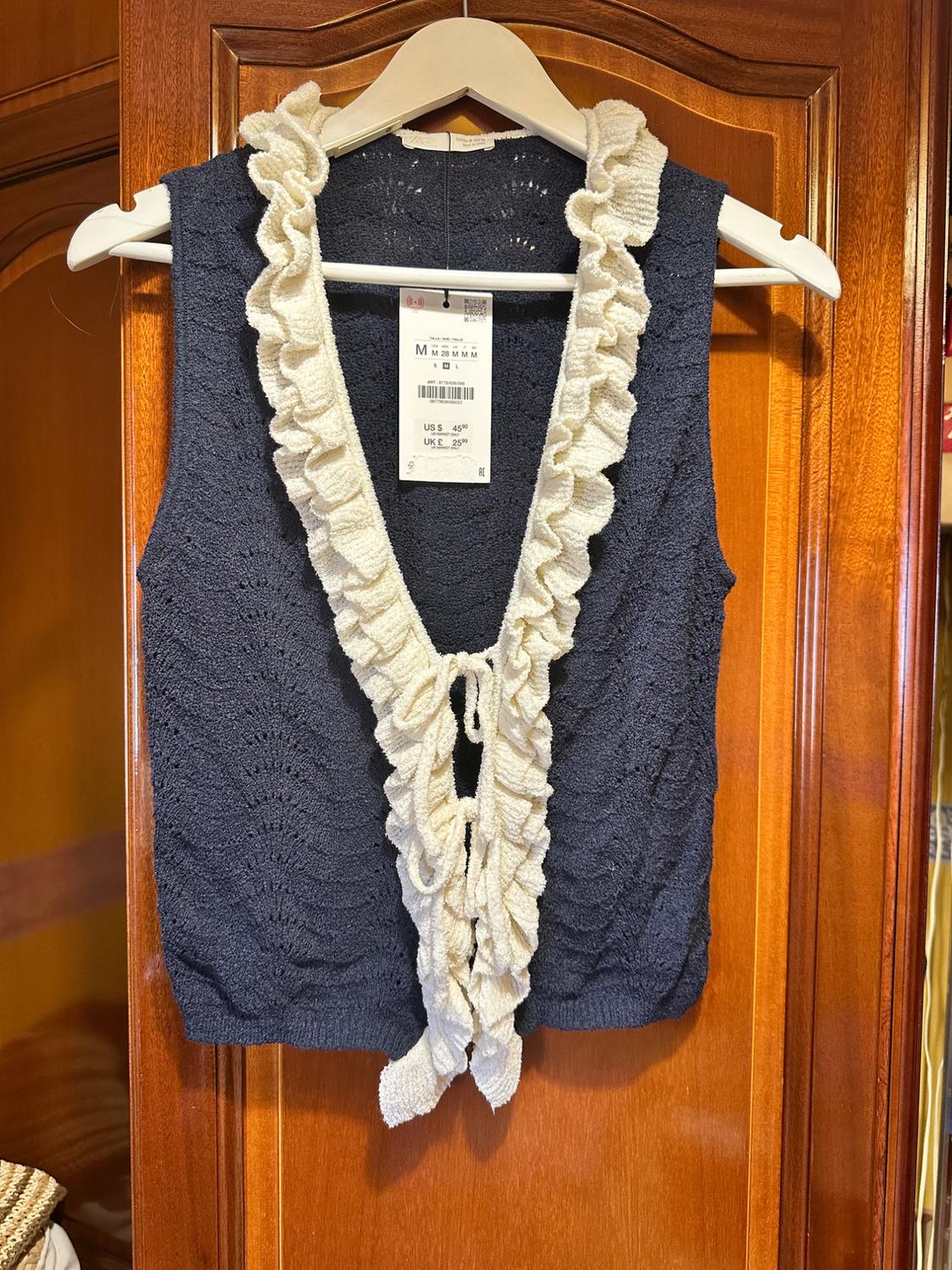 Navy knitted waistcoat with white ruffles and front tie, hanging on a white hanger over a wooden door with a visible tag.