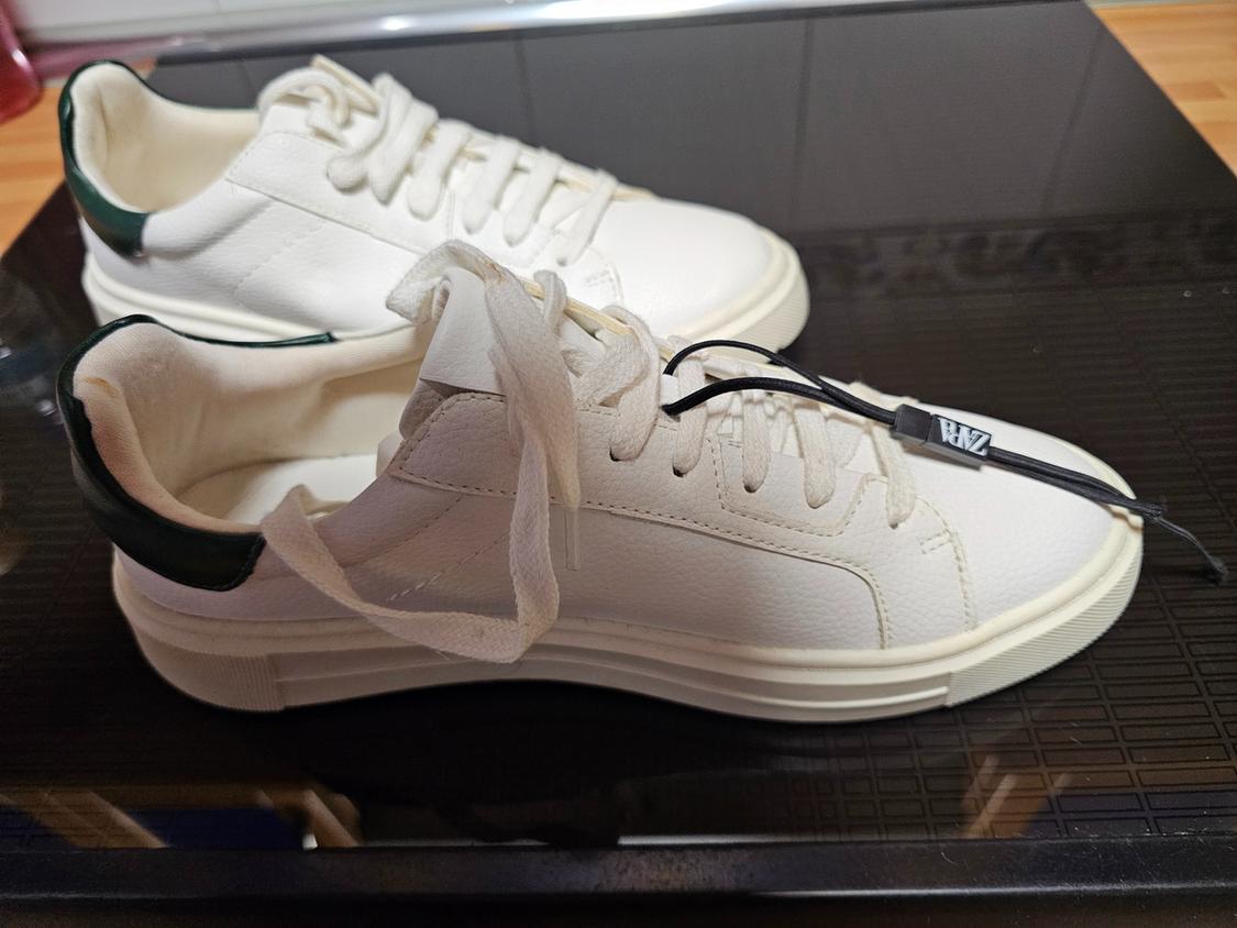 Λευκά sneakers με σκούρες πράσινες λεπτομέρειες στο πίσω μέρος και λευστά κορδόνια, το ένα με μια μαύρη ετικέτα που γράφει ZARA, πάνω σε μια σκούρα επιφάνεια.