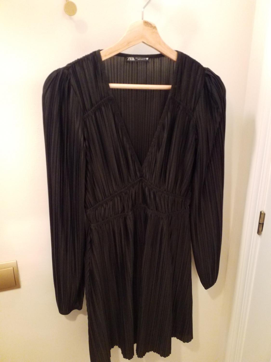 Black pleated dress with long sleeves hanging on a wooden hanger. Visible label with text "ZARA". White background.
