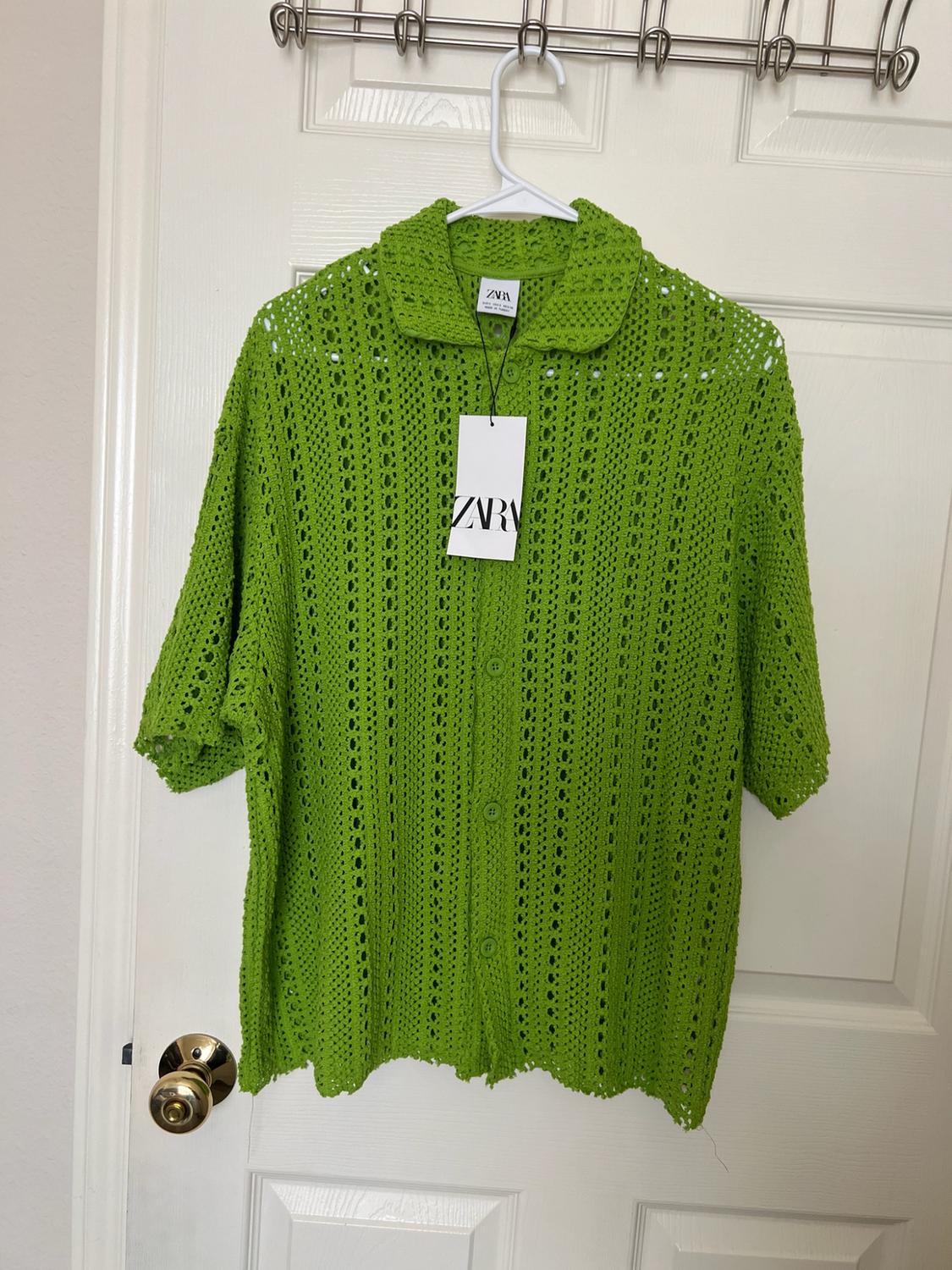 Green open-knit short-sleeve button-up shirt, hanging on a white door. Tag with the text Zara.