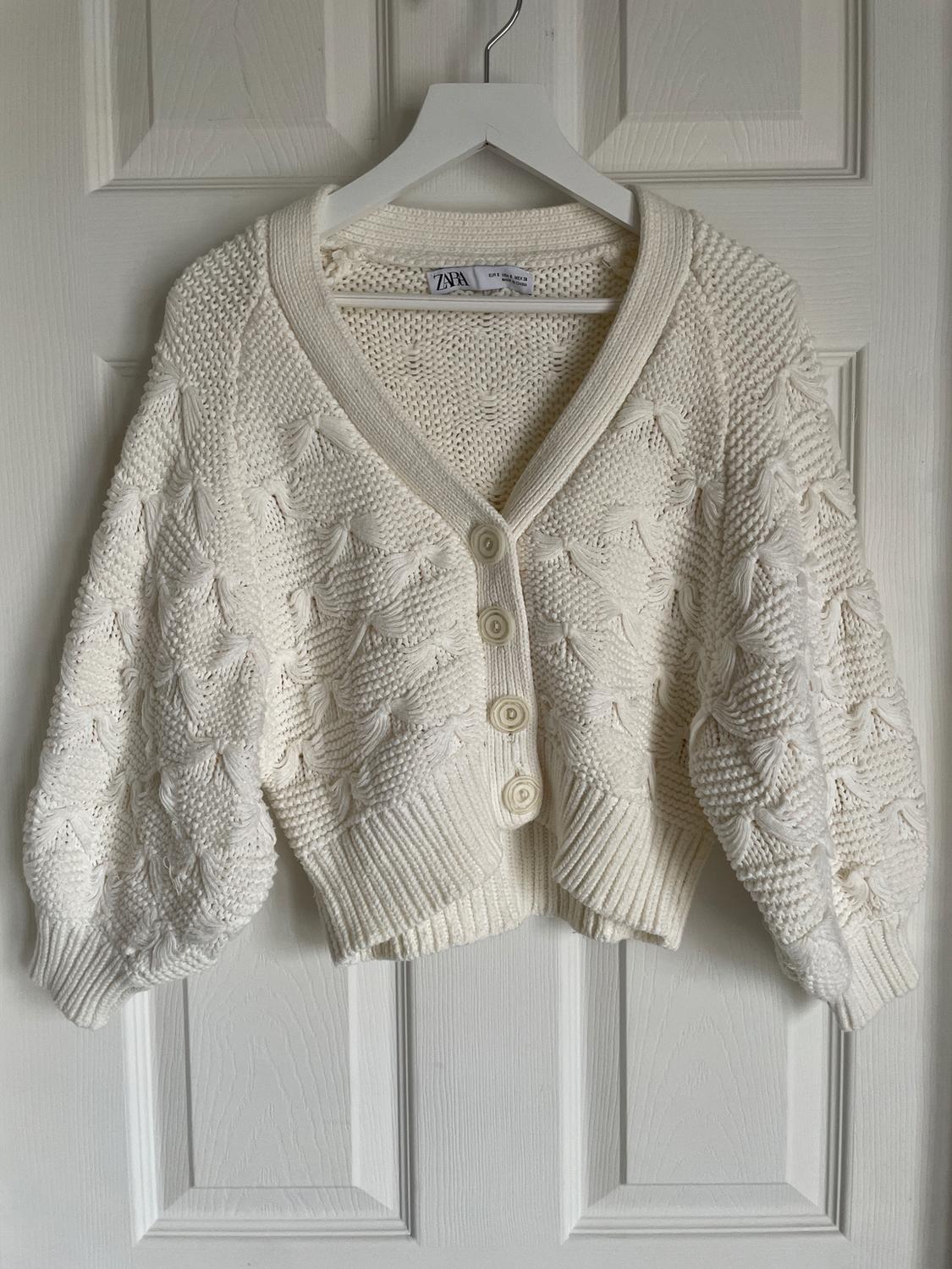 White knitted cardigan with large buttons, hanging on a white hanger in front of a white door. Label visible.