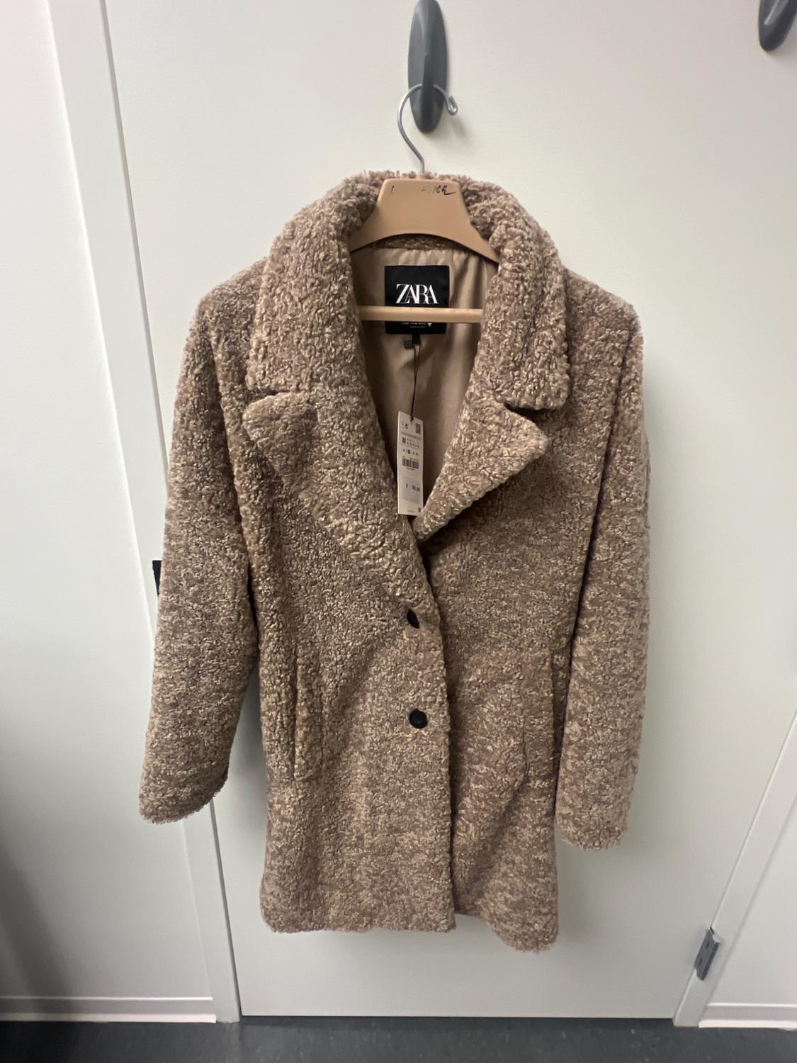 Brown wool coat hanging on a hanger with a visible Zara tag, over a white door.
