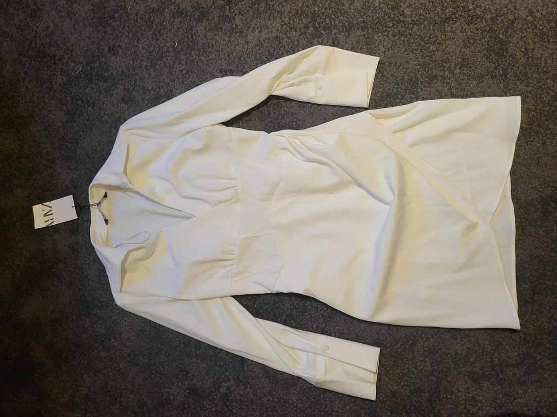 Long-sleeved white dress with a V-neck, laid on a grey surface. Visible label with text "ZARA".