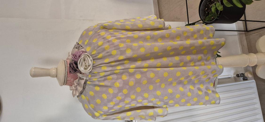 Mannequin in a yellow polka dot dress against a white background, decorated with fabric flowers at the neck. Indoor setting with a plant in the background.