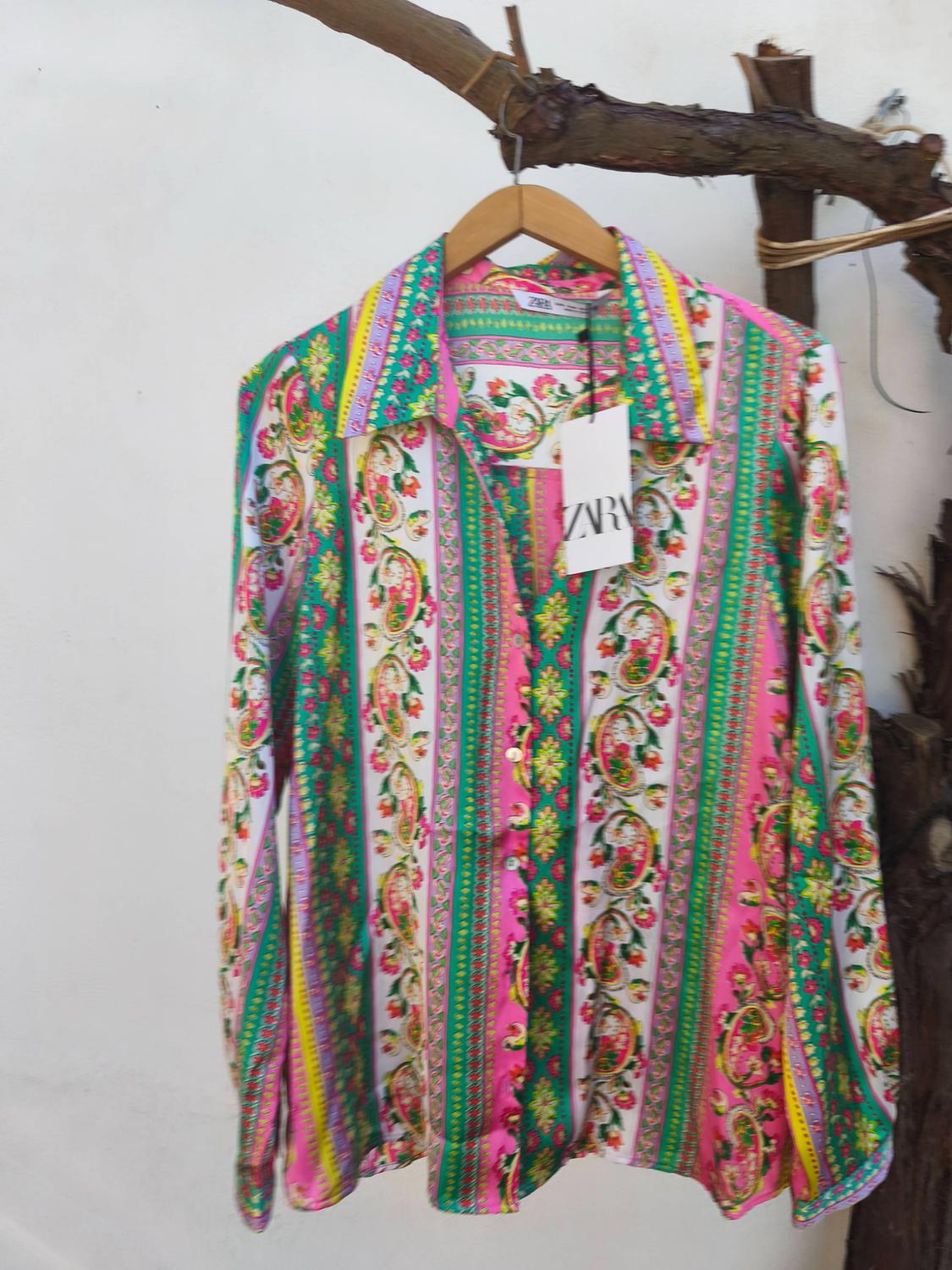 Colourful shirt with a floral print and vertical stripes in green, pink, and yellow, hanging on a wooden hanger. "ZARA" label.