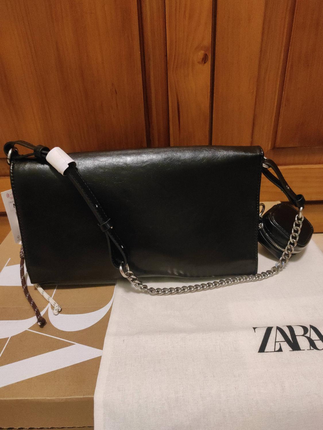 Black handbag with a silver chain and purse, on a box and a white case bearing the text ZARA, against a wooden background.