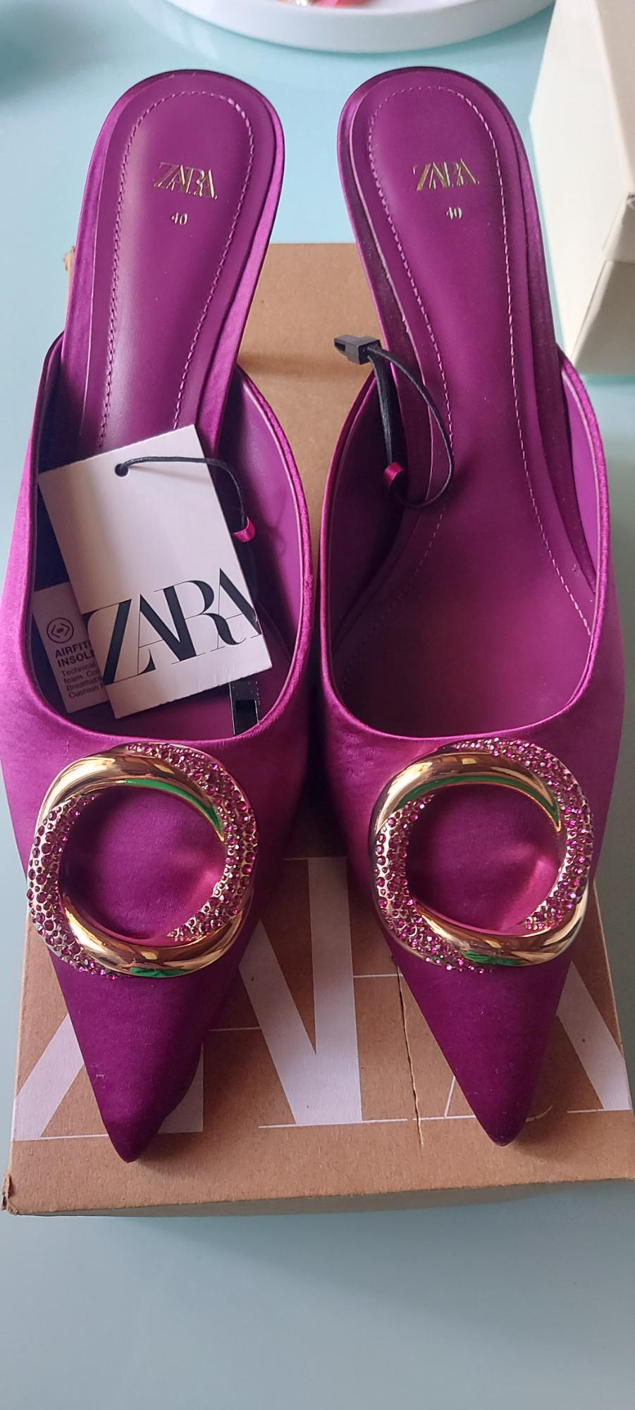 Purple pointed shoes with gold buckles and glitter details, on a cardboard box. Visible label with the text "ZARA".