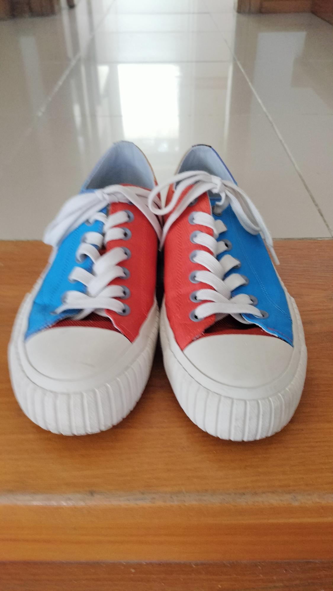 Red and blue canvas trainers with white laces and soles, on a wooden surface with a light tiled floor in the background.