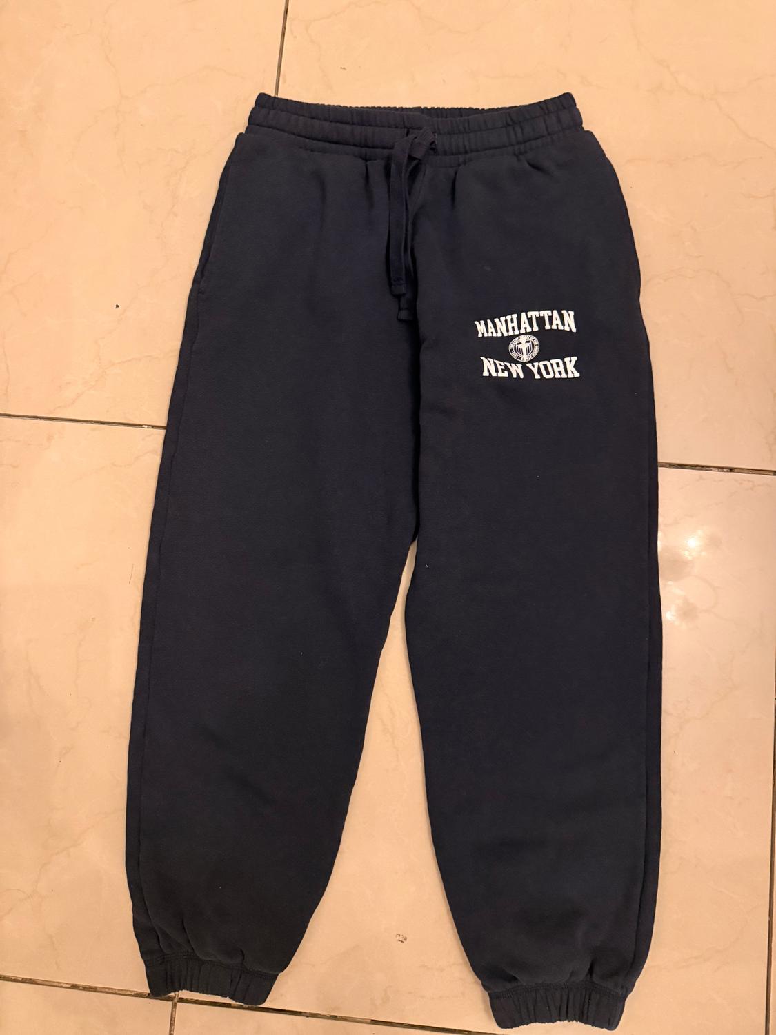 Dark blue sweatpants with elastic waist and white "MANHATTAN NEW YORK" text on the left leg, on a beige floor.