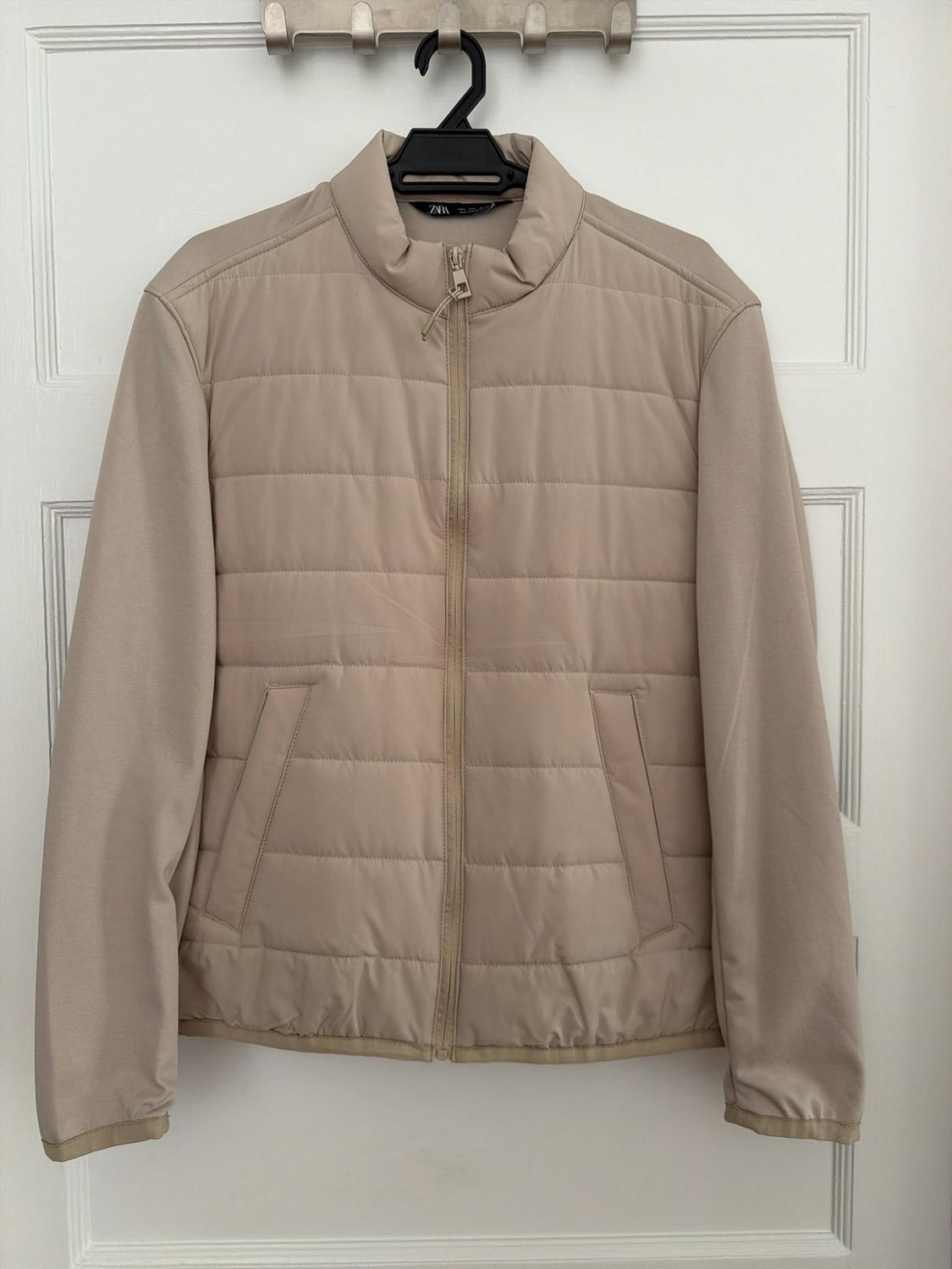 Beige jacket with quilted body, smooth sleeves, zipper, and ZARA label. Hanging on a black hanger against a white door.