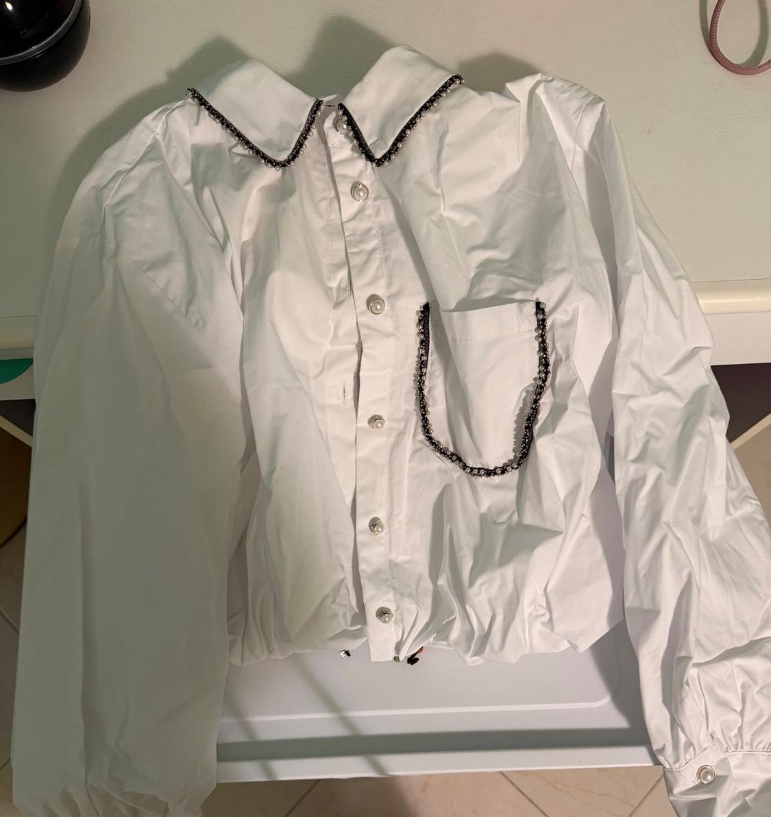 White shirt with buttons, collar, and pocket adorned with a black chain. Long sleeves and a light background.