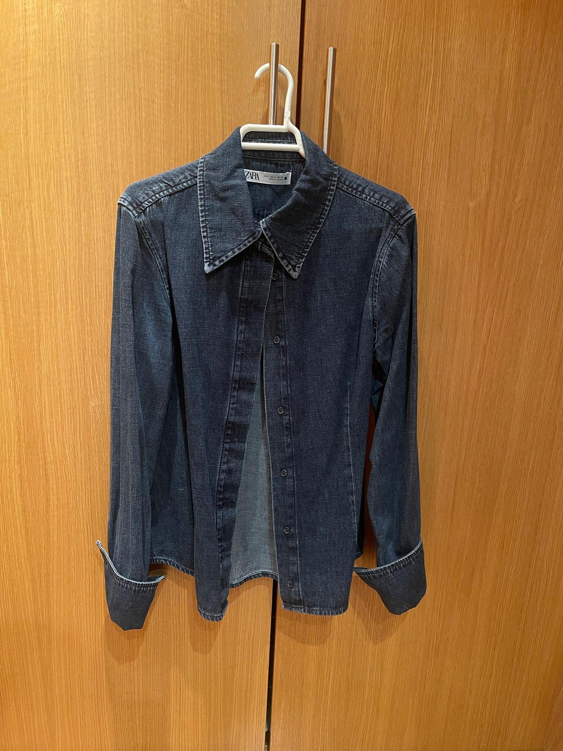 Dark blue denim shirt hanging on a white hanger in front of a light wooden door. Visible label: ZARA.