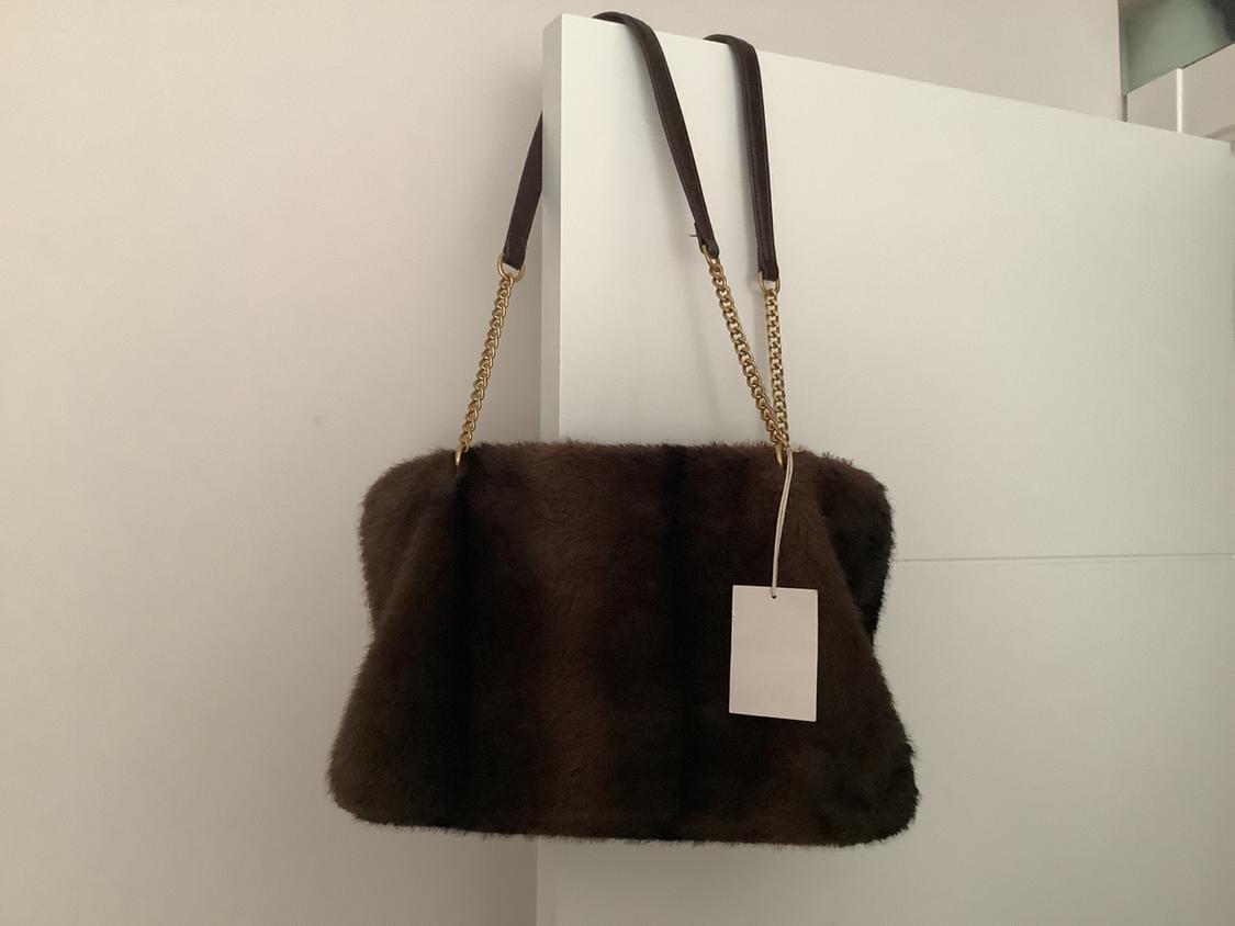 Brown fluffy-textured handbag with a gold chain and a plain white tag. It hangs from a white door in a relaxed and minimalist setting.