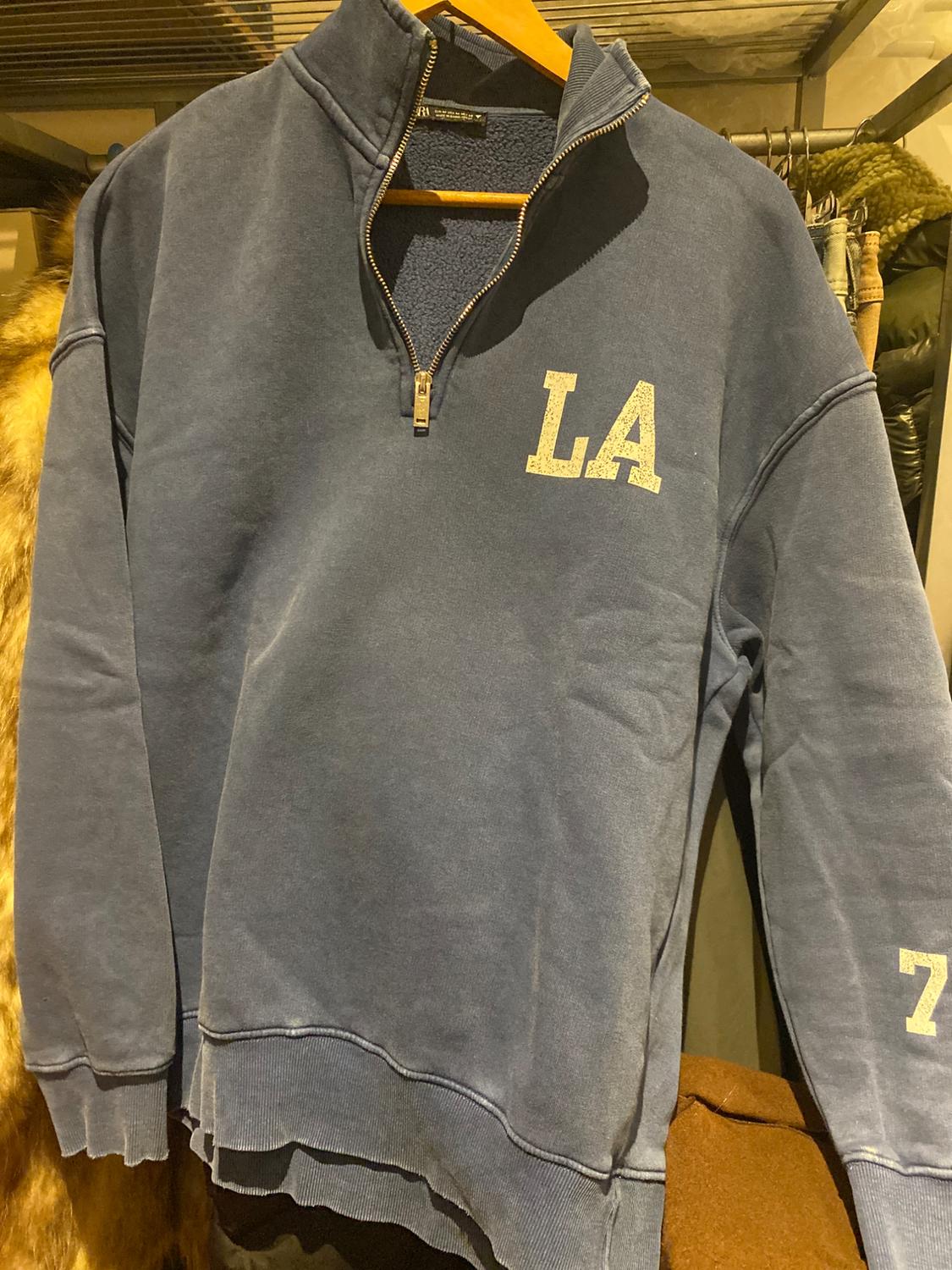 Blue sweatshirt with a quarter-zip, hanging on a hanger. "LA" lettering on the chest and number "7" on the sleeve.