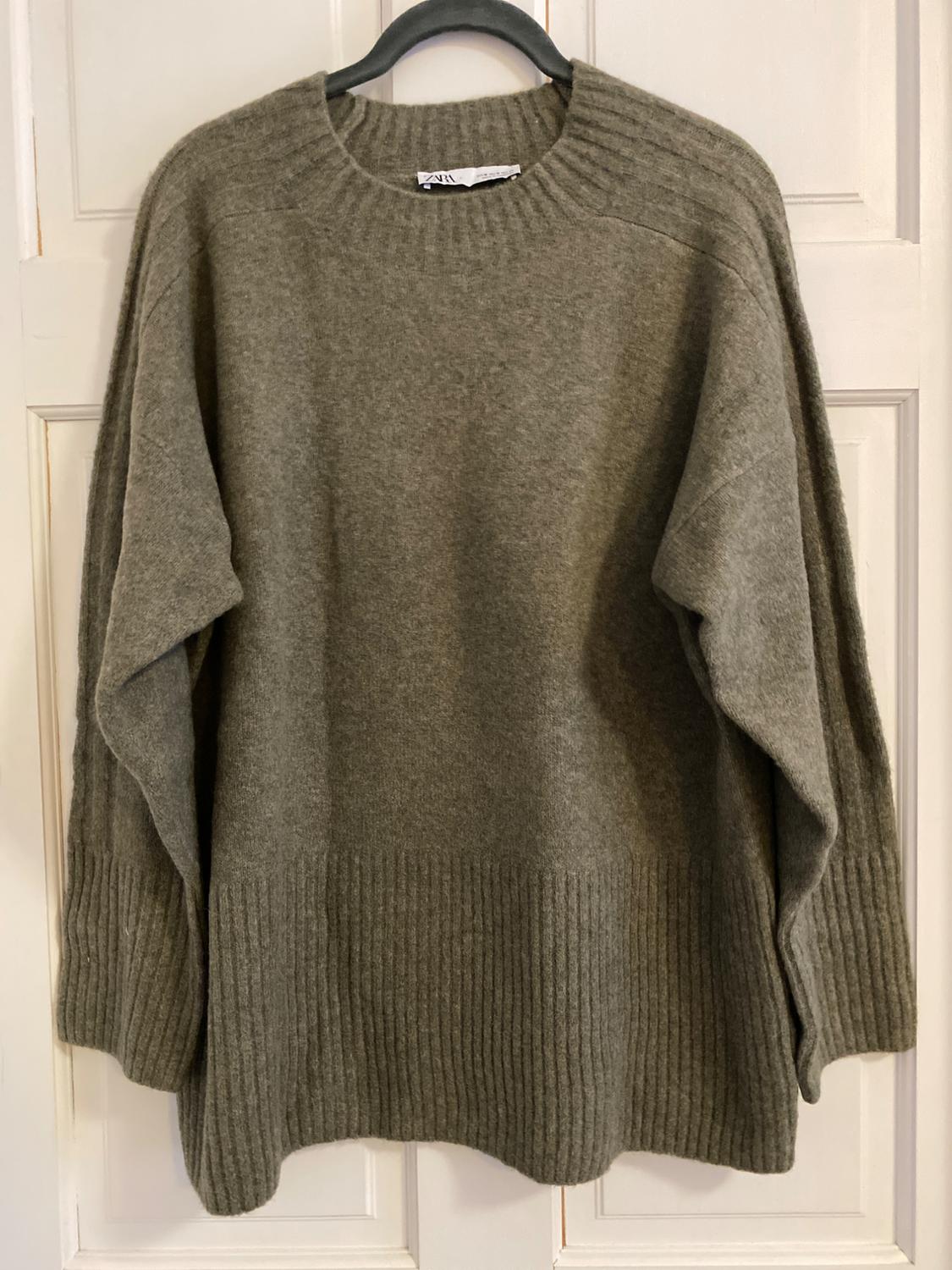 Gray chunky knit sweater hanging on a black hanger over a white door. Crew neck design with long sleeves.