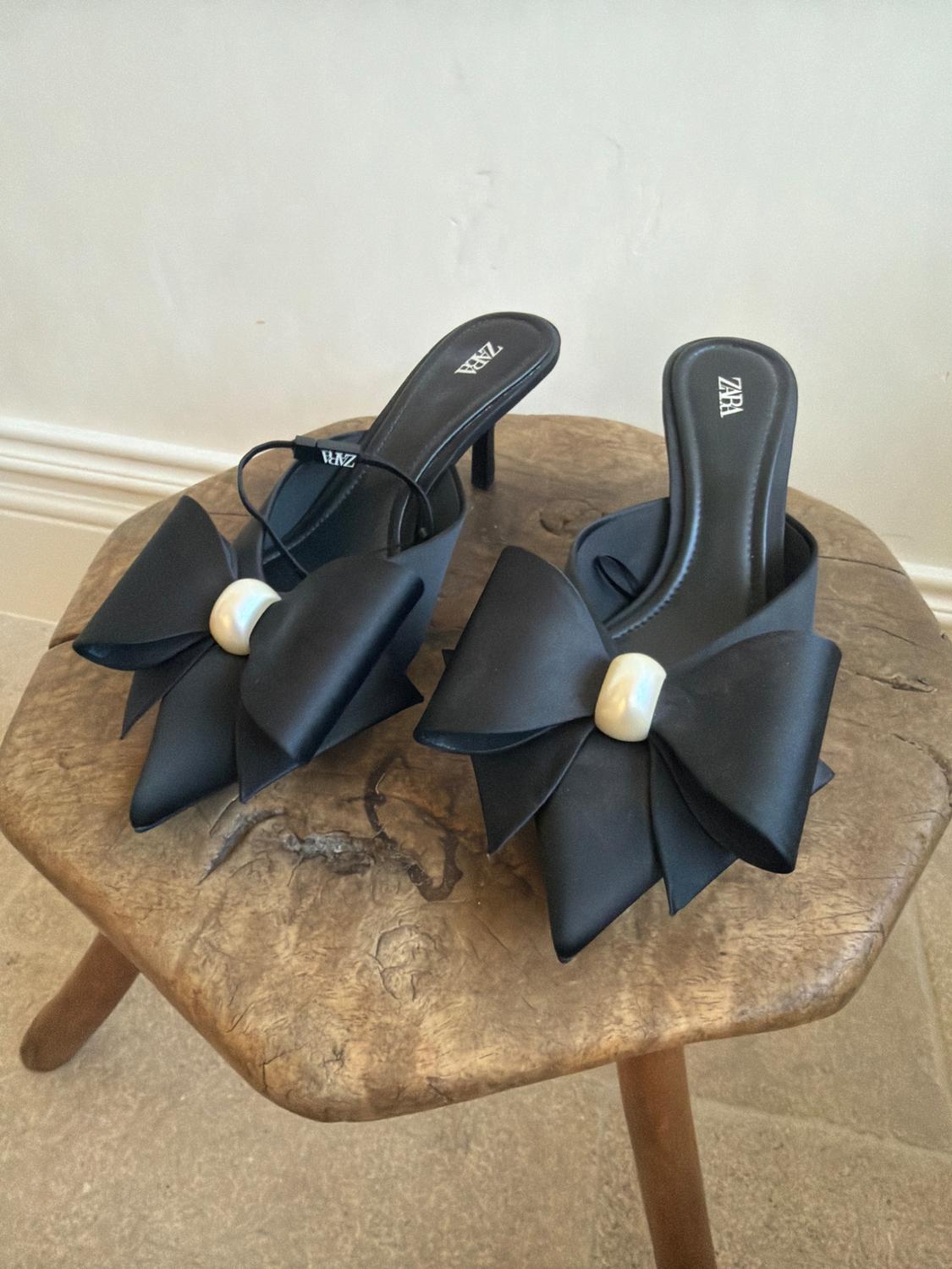 Black pointed-toe high heels with a large bow and pearl on a rustic wooden stool. The insole reads ZARA.