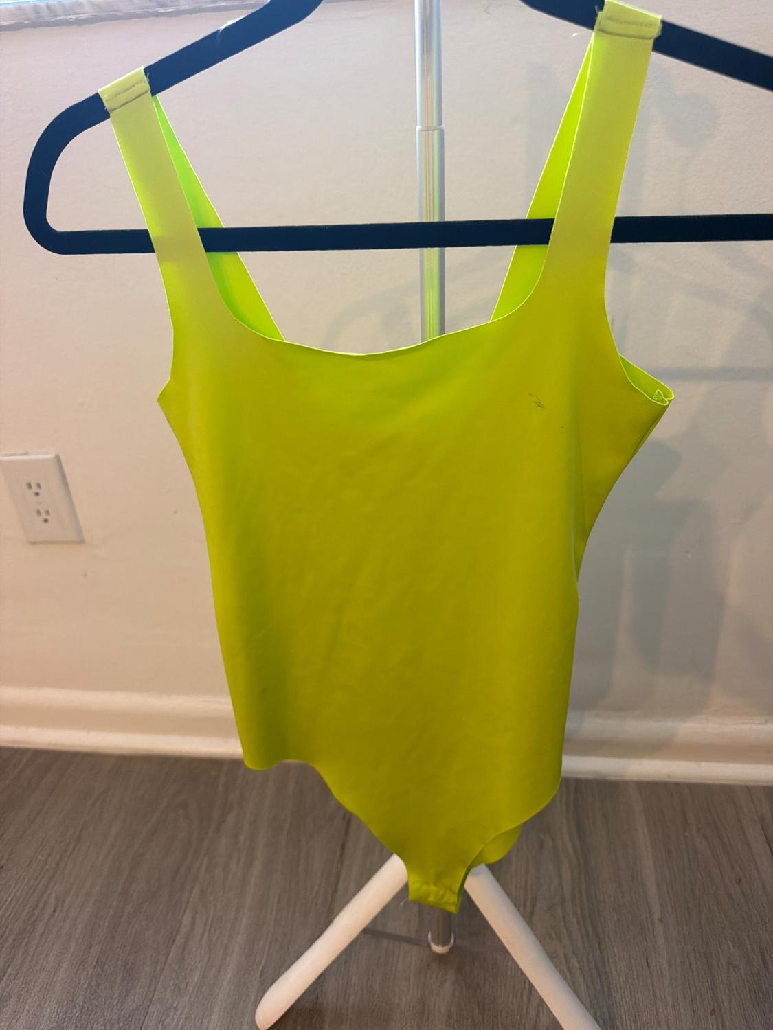 Neon yellow bodysuit hanging on a black hanger on a white garment rack. White wall background with an electrical outlet and a wooden floor.