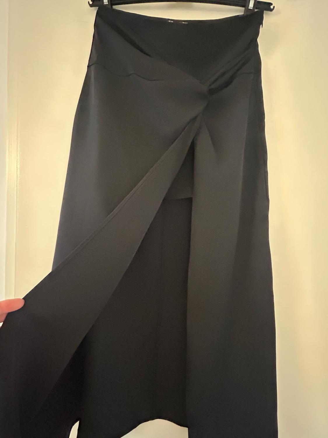 A long black skirt with a wrap-around design, hanging on a hanger. A hand holds the edge of the fabric, showing how it drapes.