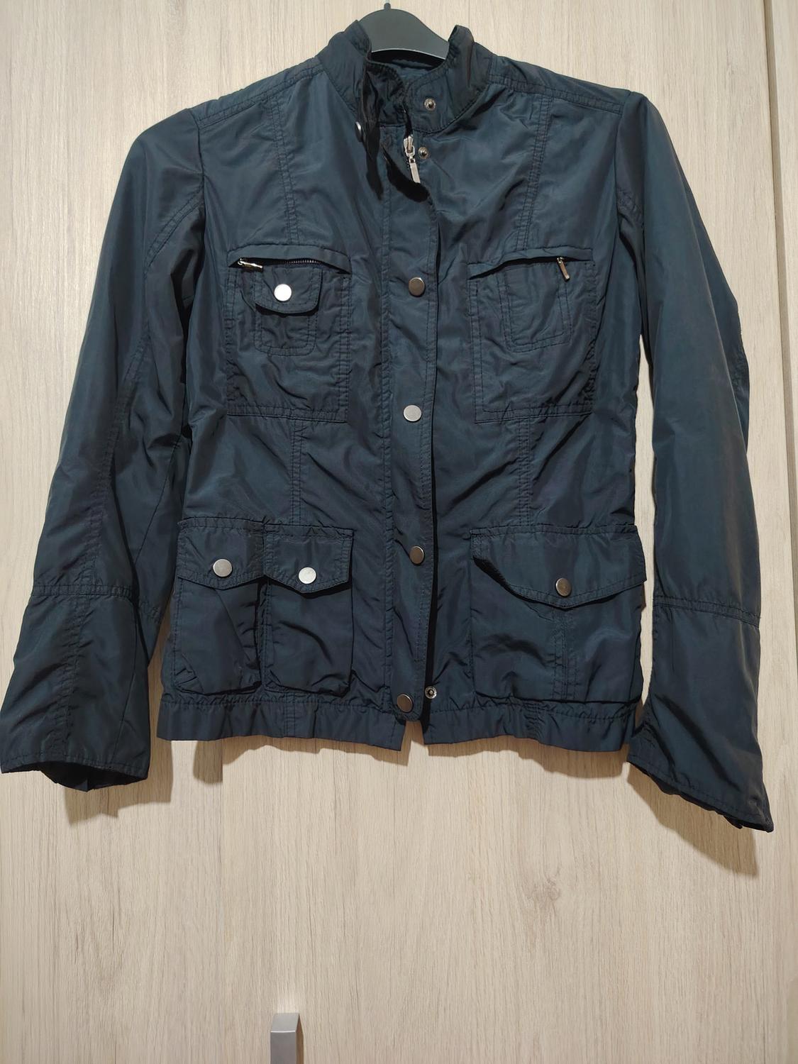 Black jacket with multiple pockets and silver buttons, hanging on a hanger against a light wooden background.