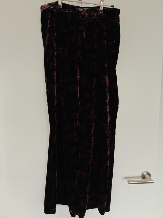 Black velvet trousers with a dark red floral print, hung on a hanger against a white background.