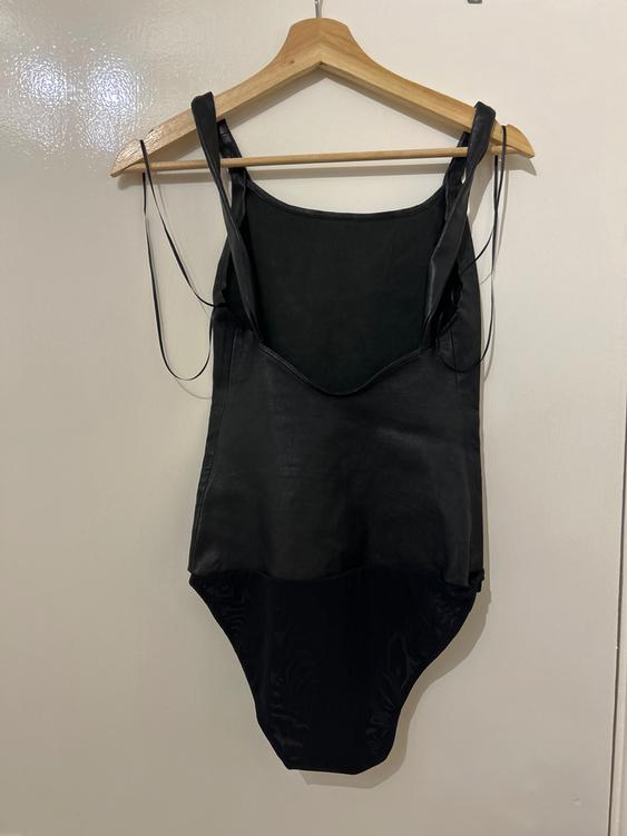 Black bodysuit hanging on a wooden hanger against a white wall. Simple design with thin straps and a round neckline.