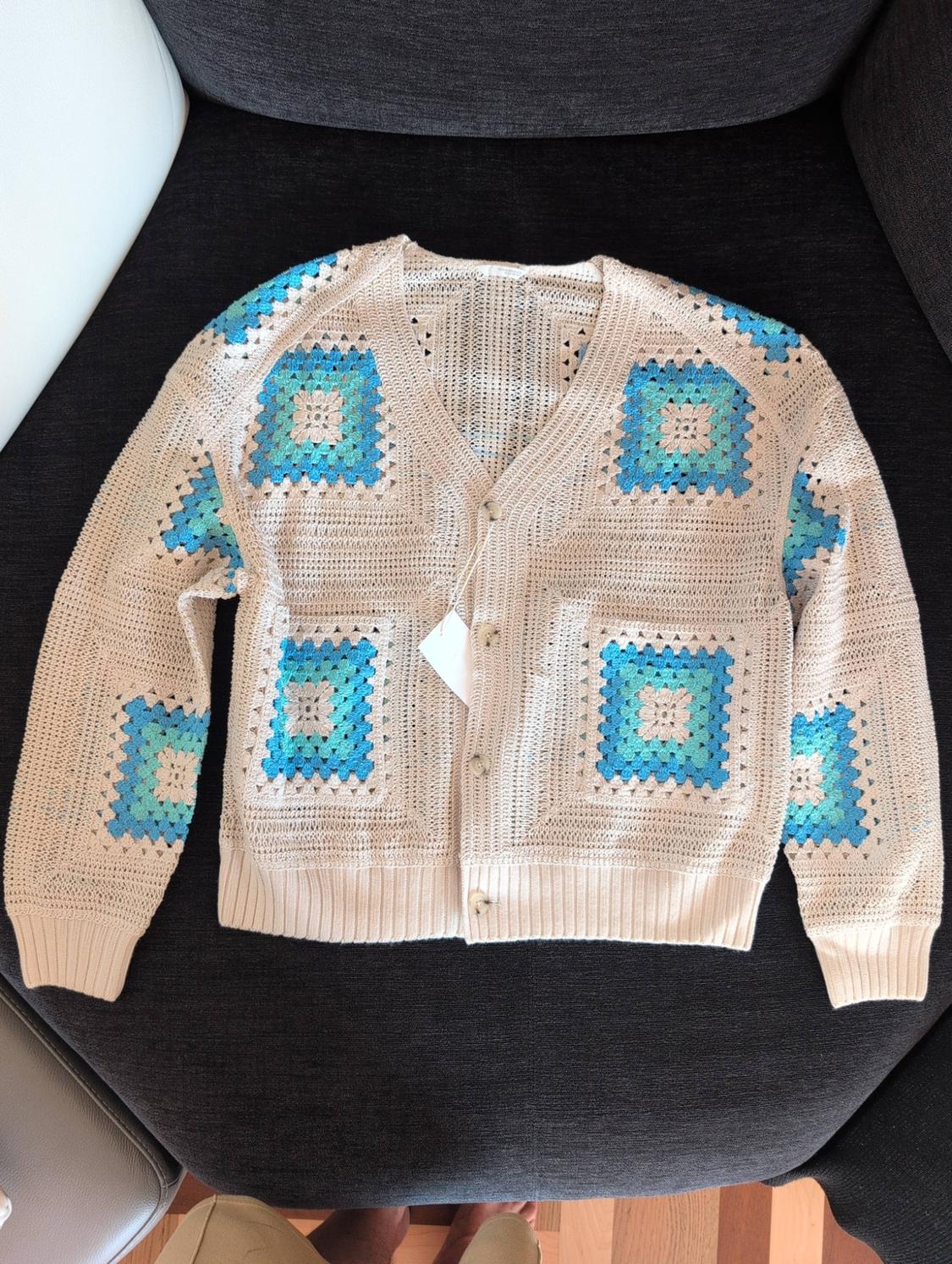 Beige knitted cardigan with blue and turquoise squares, front buttons and label, on a dark sofa.