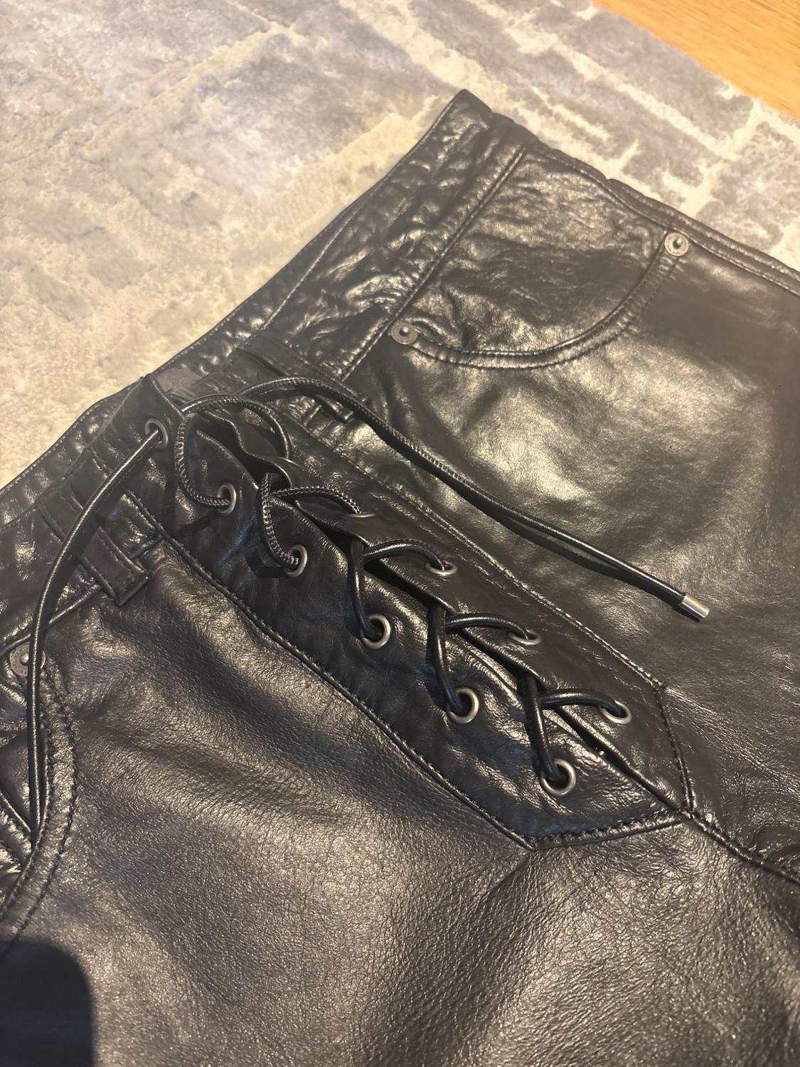 Black leather trousers with lace-up detail on the side, on a grey rug with a geometric pattern.