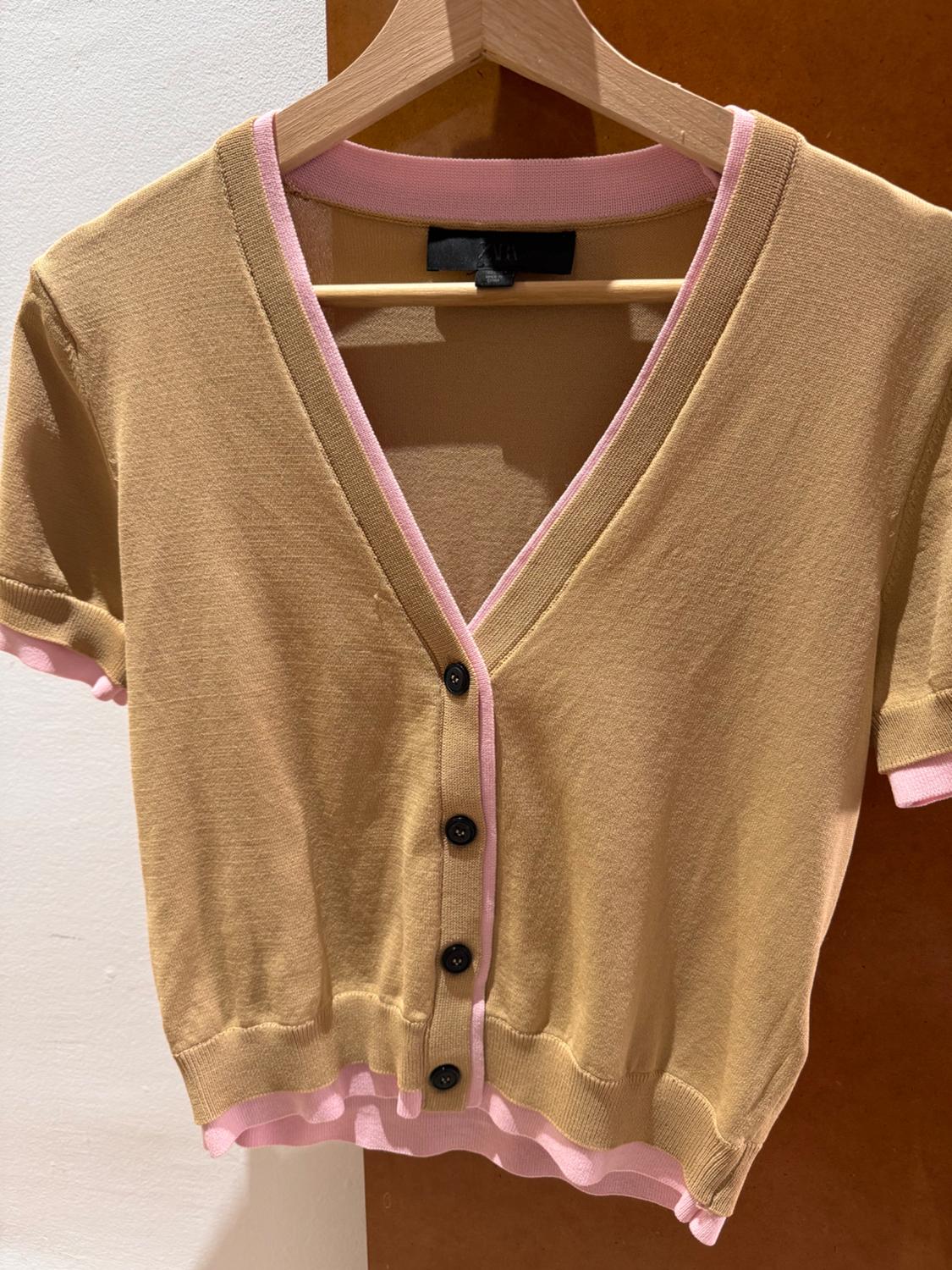 Short beige cardigan with pink trim and black buttons, hanging on a wooden hanger against a brown background.