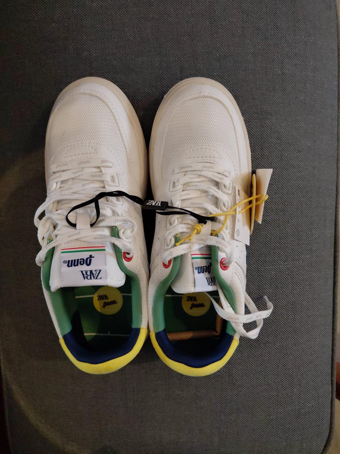 White trainers on a grey background with green, blue and yellow details. They feature the text ZARA and “penn” on the tongue, laces and insole.