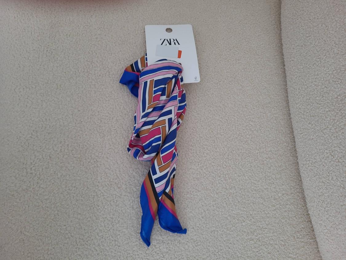 Colourful scarf with a geometric print in shades of blue, pink, and brown, on a beige background. Visible label with "ZARA" logo.