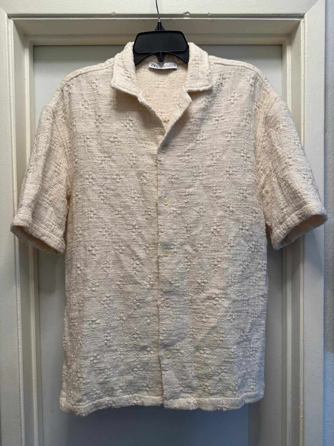 Cream-colored textured fabric shirt, short-sleeved and open-collared, hanging on a black hanger on a white door.